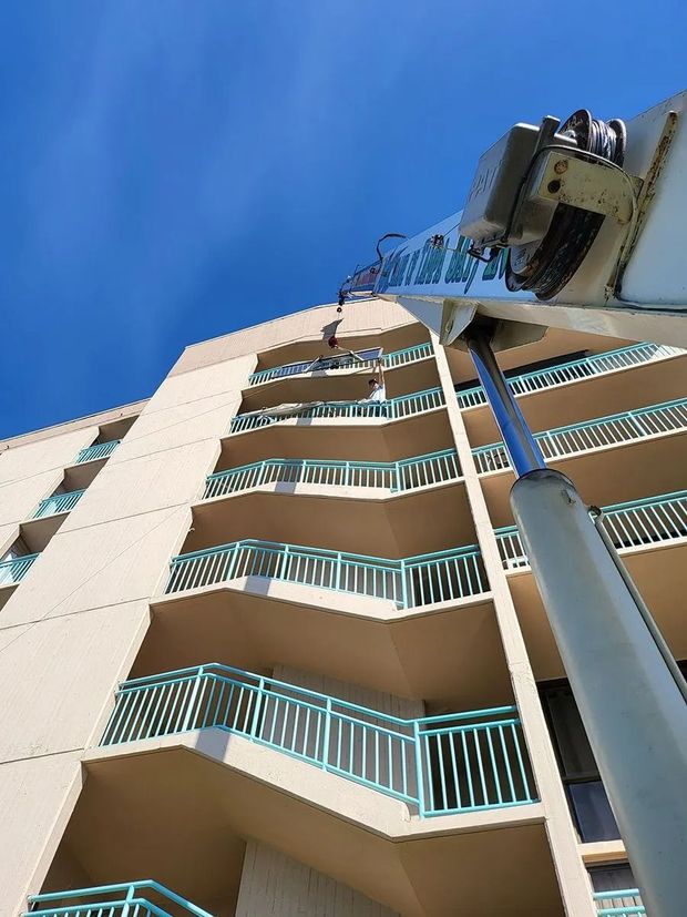 A crane arm reaches toward the balconies of a tan, multi-story building against a clear blue sky.