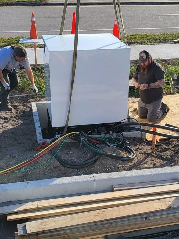 Two workers install a large, white electrical cabinet on a concrete pad near a roadside, guided by overhead lifting straps.