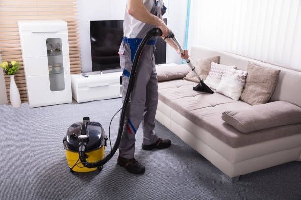 A man is using a vacuum cleaner to clean a couch in a living room.