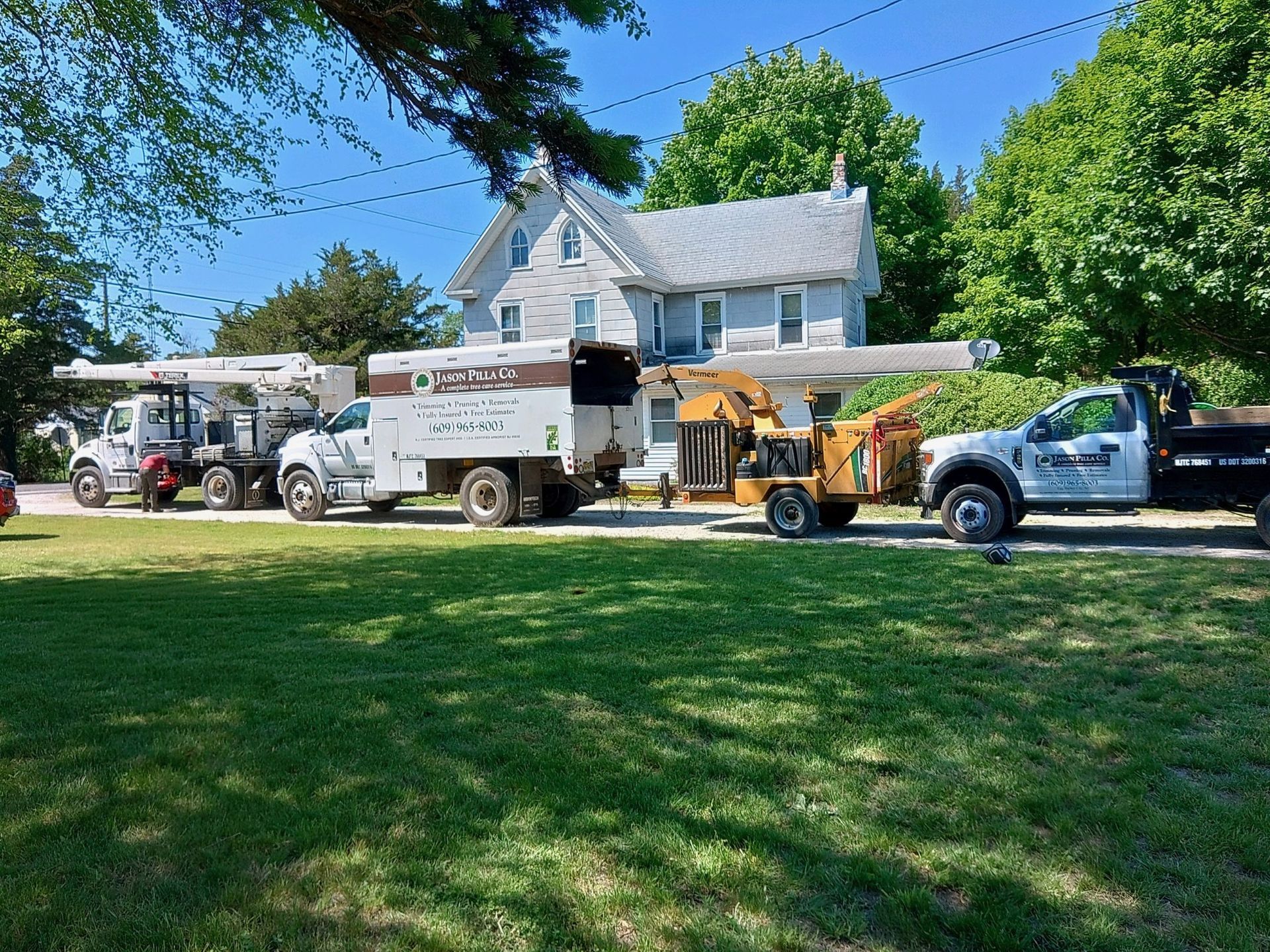 Tree service trucks and equipment parked in front of a gray house, on a sunny day.