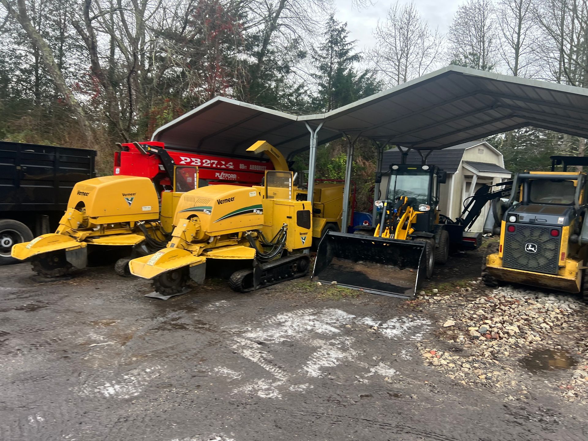 Heavy machinery parked under a metal canopy. Includes stump grinders, a wood chipper, and a skid steer, outside.