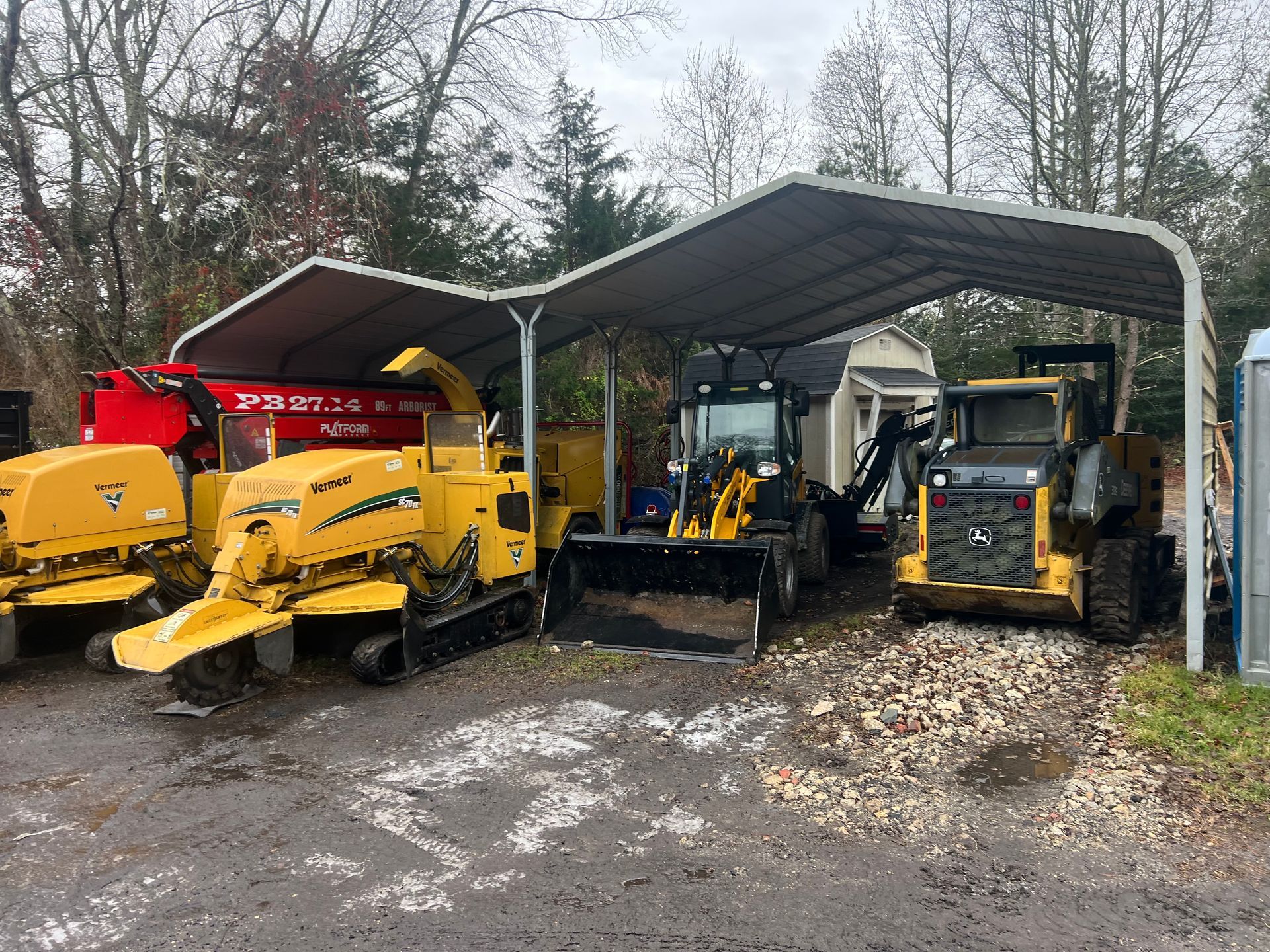 Equipment under a carport: chipper, stump grinders, front-end loader, and skid steer.