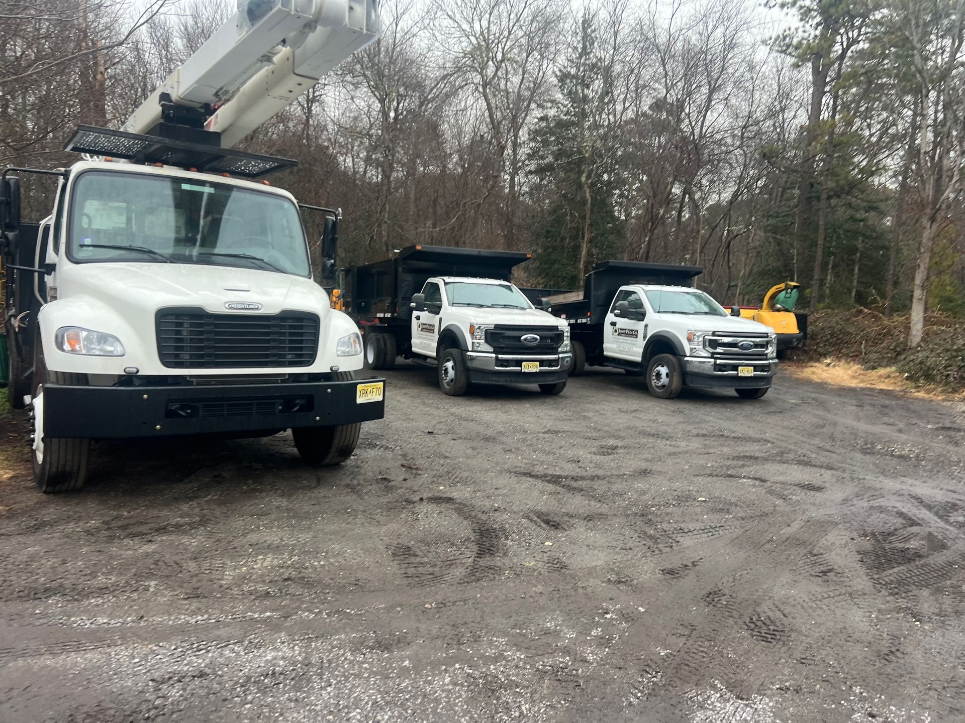 White trucks parked on a muddy lot, including a boom truck and dump trucks. Forest in the background.