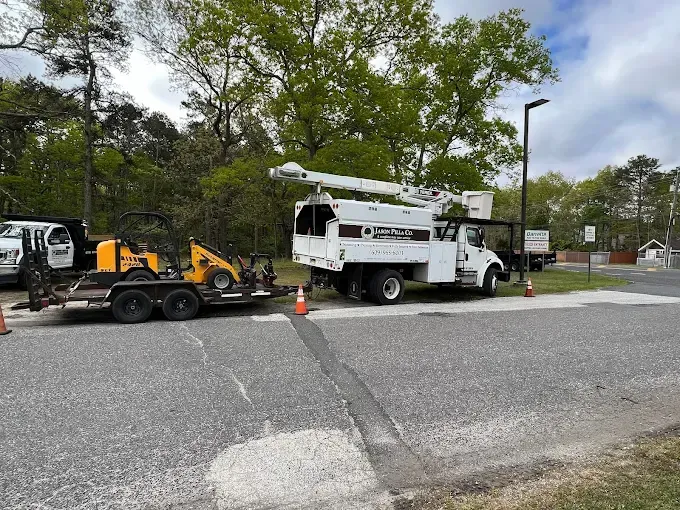 White bucket truck and trailer with equipment parked on asphalt near trees.