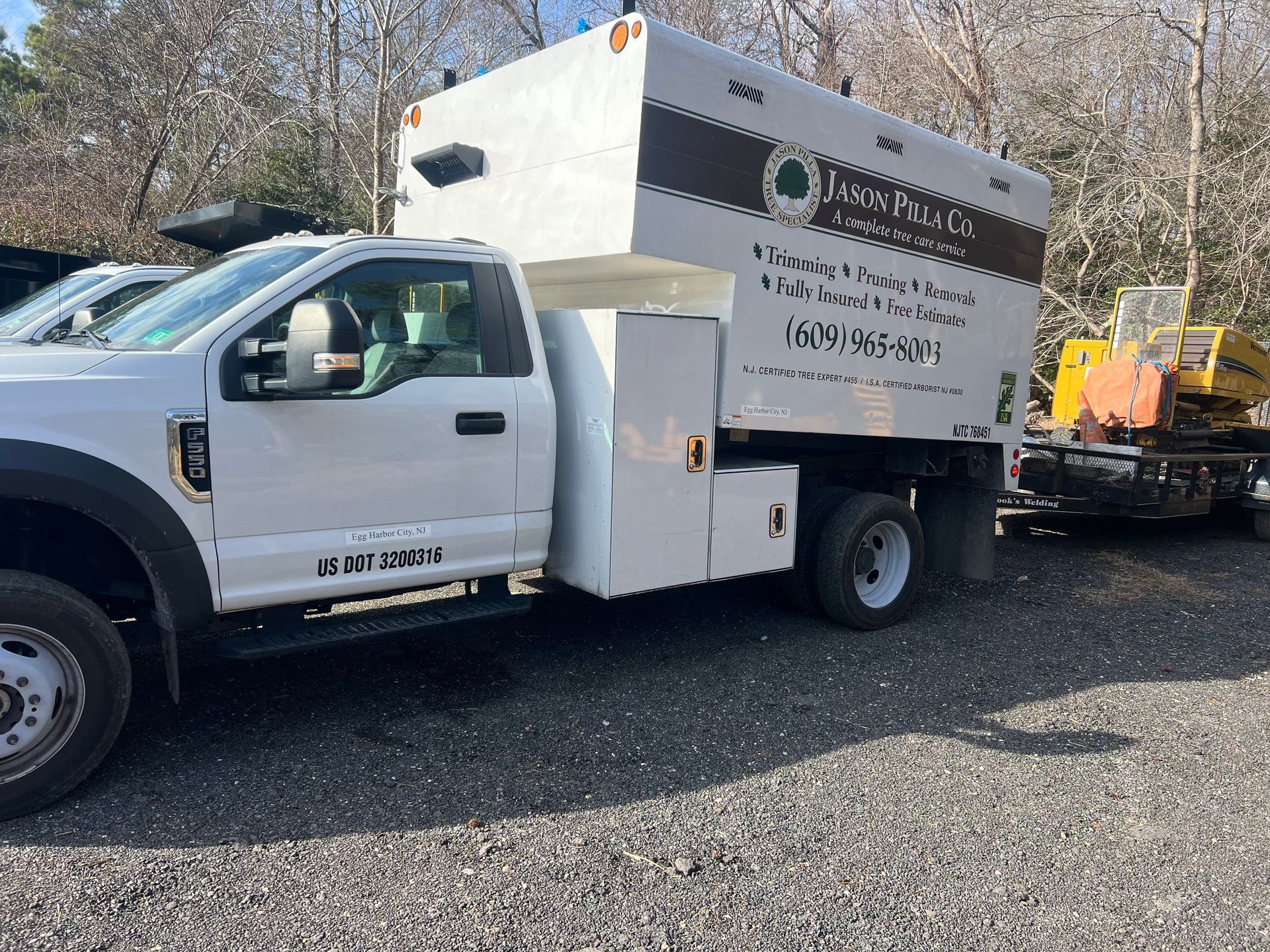 White tree service truck parked on gravel, logo on side, yellow construction equipment in background.