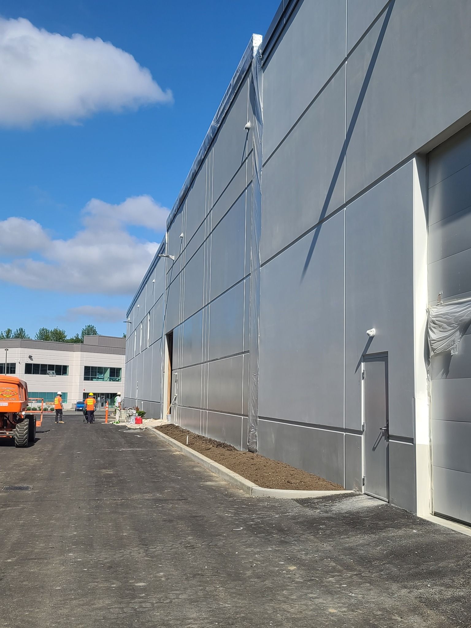 Exterior of a modern industrial building, silver metal siding, partially open door, and construction workers visible.