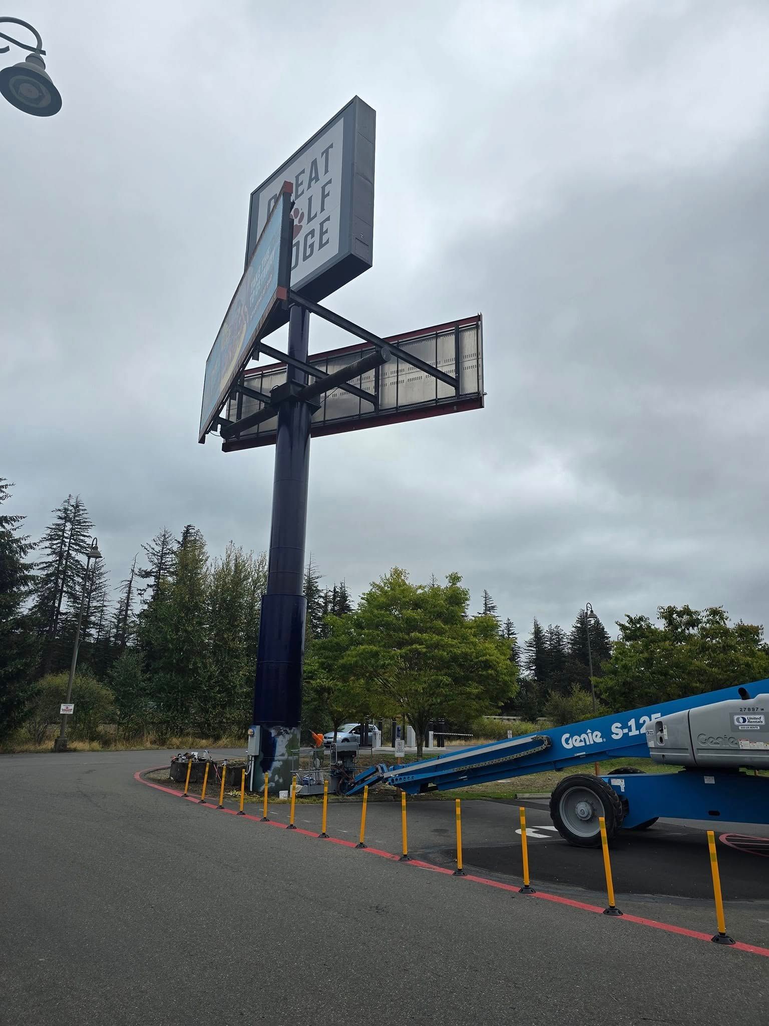 Billboard being worked on, blue lift in parking lot. Cloudy sky, trees in background.