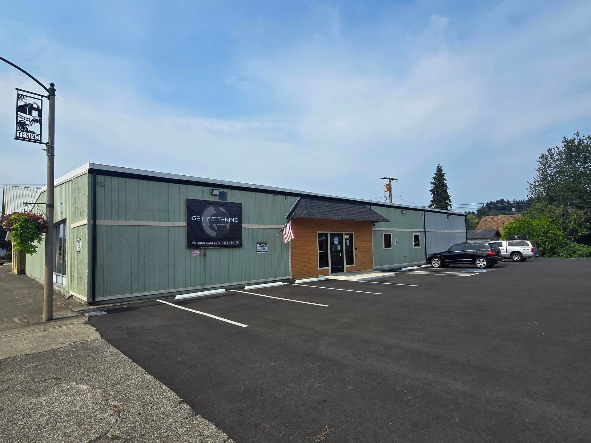 Building with a black sign above a brown door, American flag, parked cars, blue sky.