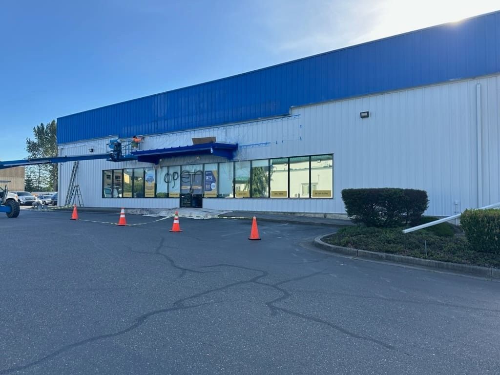 Exterior of a business with blue and white facade, large windows, orange traffic cones, and overcast sky.