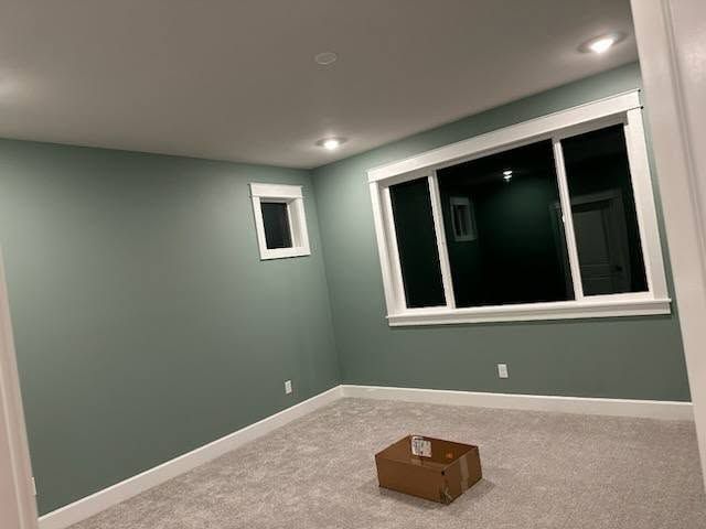 Empty room with green walls, white trim, recessed lighting, and a large window. A cardboard box sits on the light gray carpet.