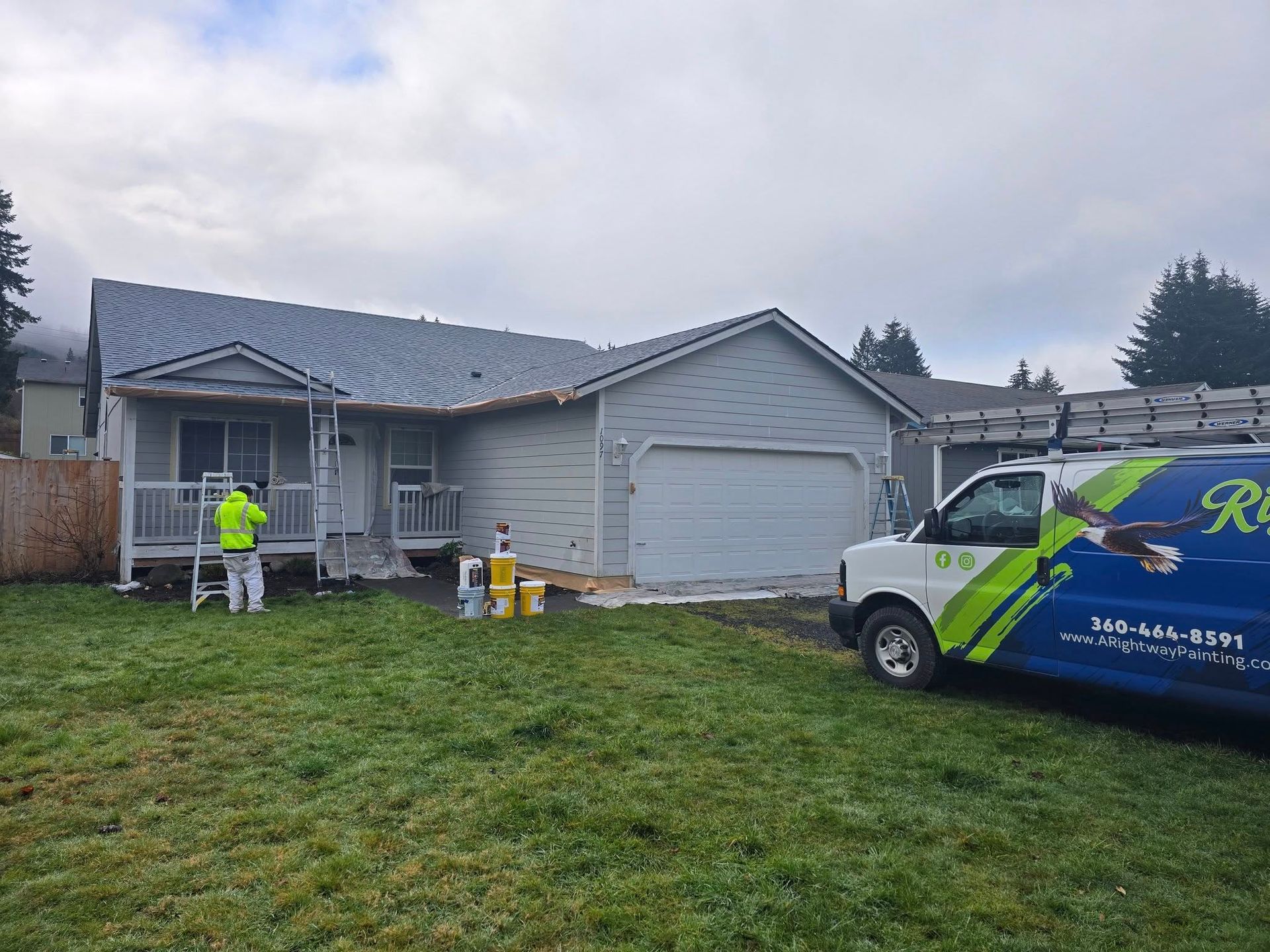 A house being painted gray with a contractor in a yellow vest and a company van on the lawn.