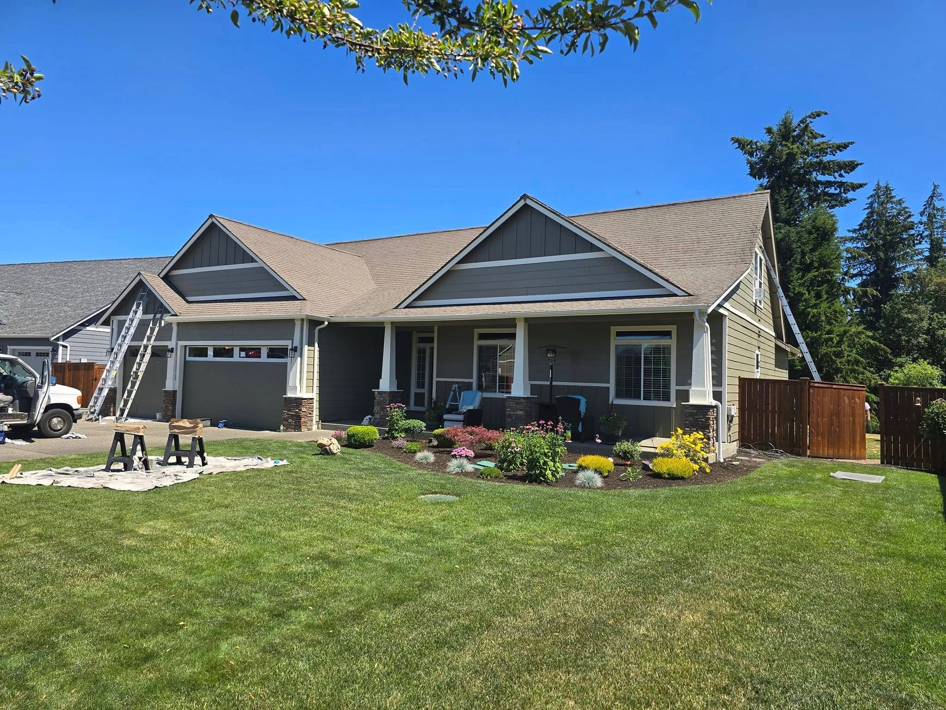 Gray house with green lawn; ladders and painting supplies visible; blue sky.