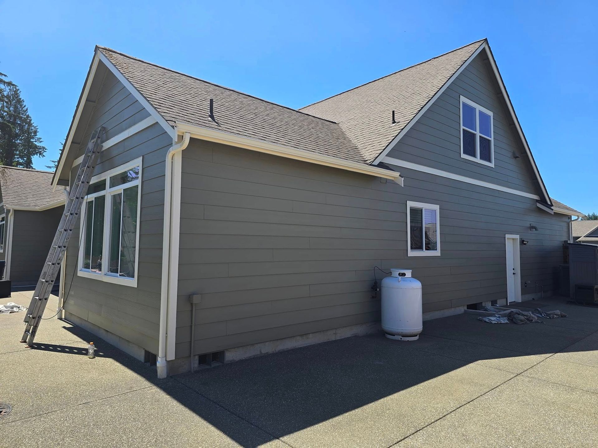Green house with white trim, gray roof, and propane tank on a concrete driveway under a blue sky.