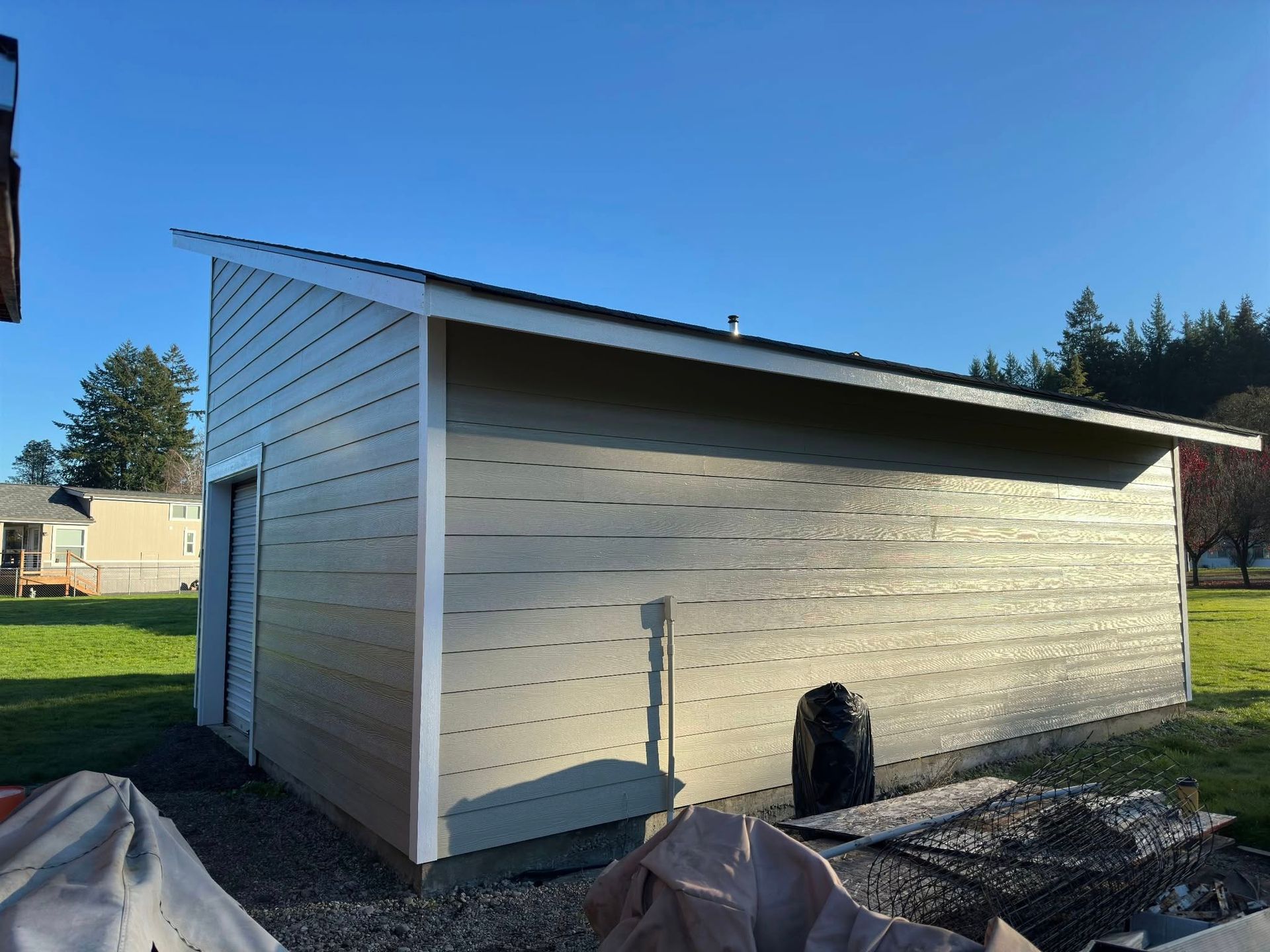 Beige shed with angled roof, white trim, and a black chimney, on a grassy lawn under a blue sky.