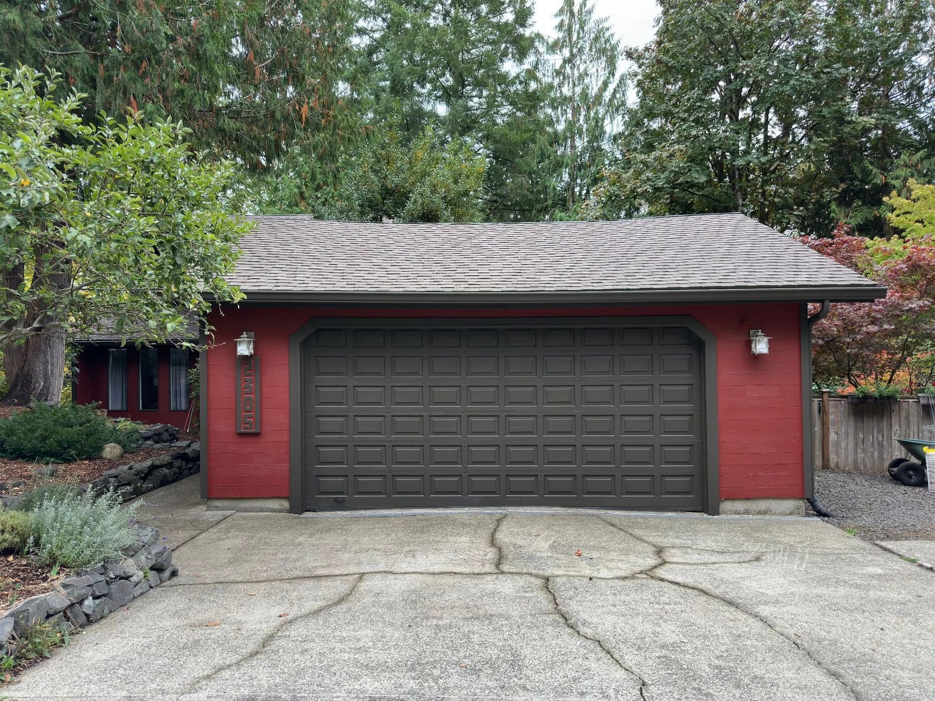 Red-painted house with a brown garage door. Concrete driveway is cracked. Green trees surround the building.