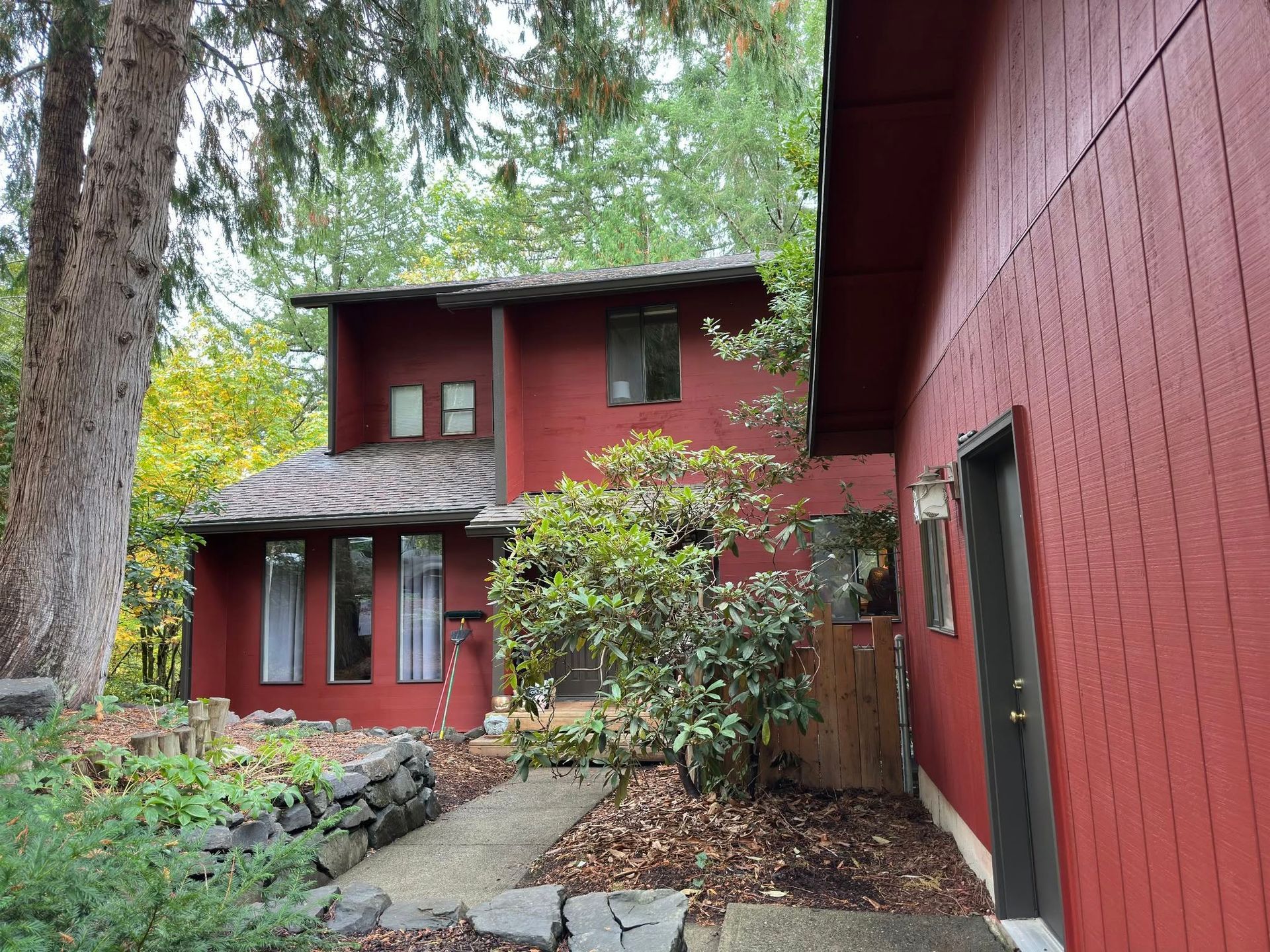 Red-sided house with multiple windows and a path leading to the entrance, surrounded by trees and foliage.