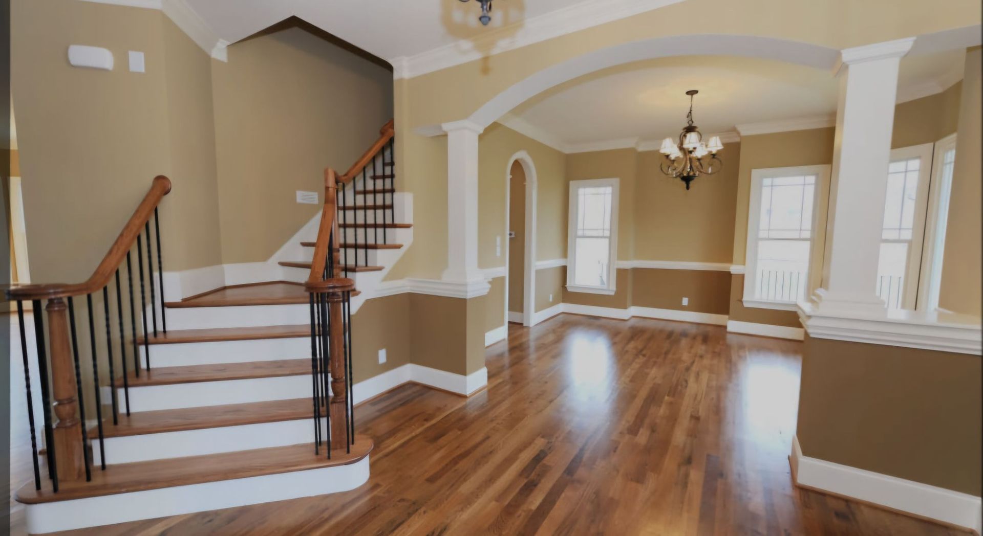 Interior view of a house with wooden floors, staircase, and dining area with arched doorway and chandelier.