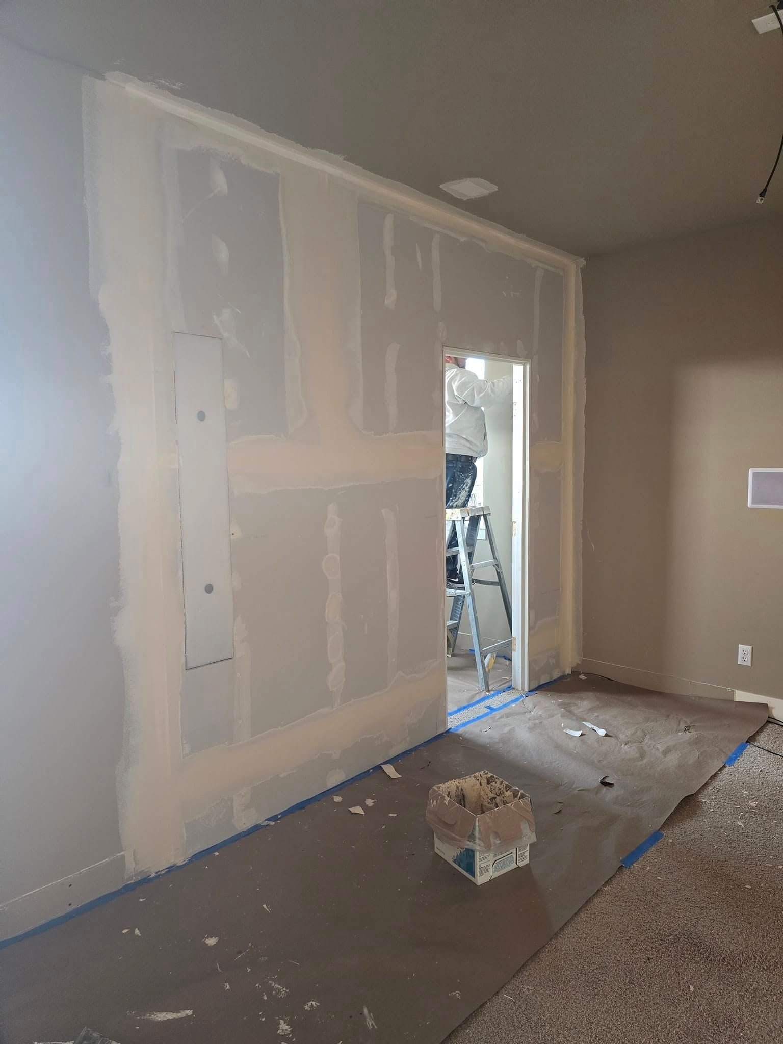 Drywall installation in progress; worker inside doorway. Wall and floor covered in mud and blue protective tape.