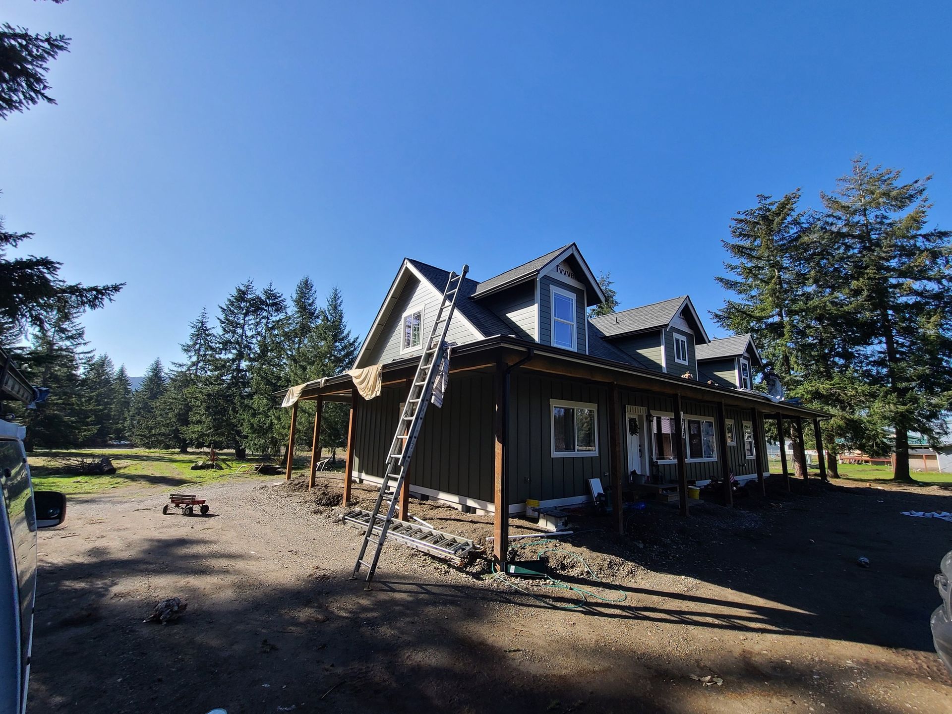 Gray house with porch under construction, ladder leaning against it, on a gravel lot under a clear blue sky.