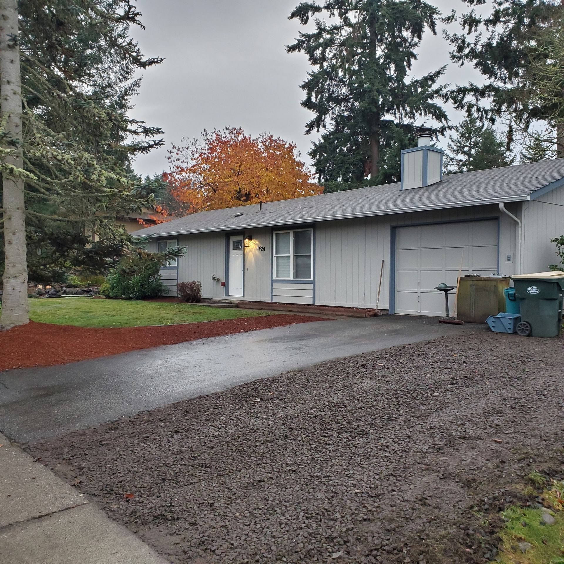 Gray house with a driveway and gravel, surrounded by trees. Cloudy day with fall foliage.