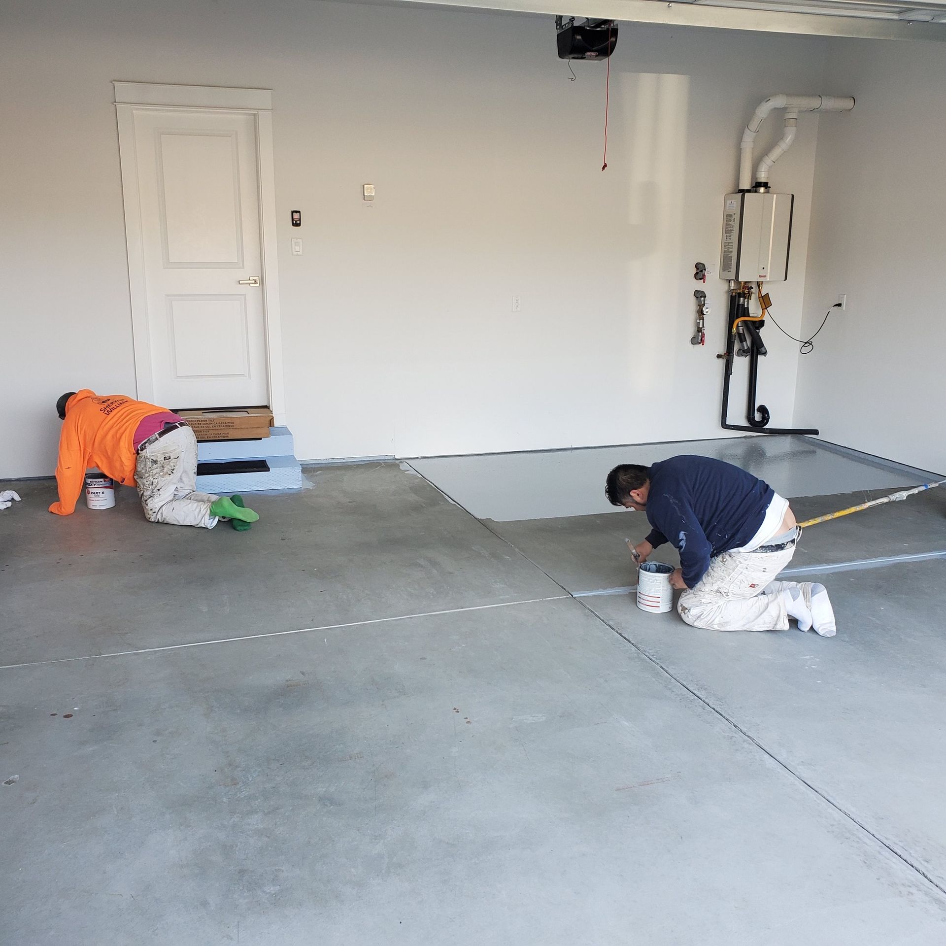 Two people painting a garage floor, kneeling. Gray concrete, white walls, and a white door.