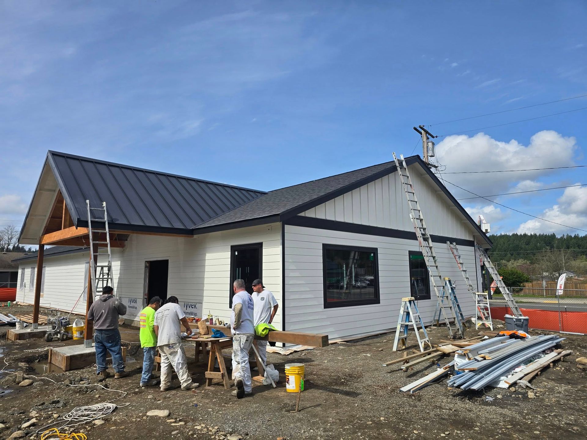Construction workers painting a white house with a black roof under a blue sky. Ladders and tools are present.