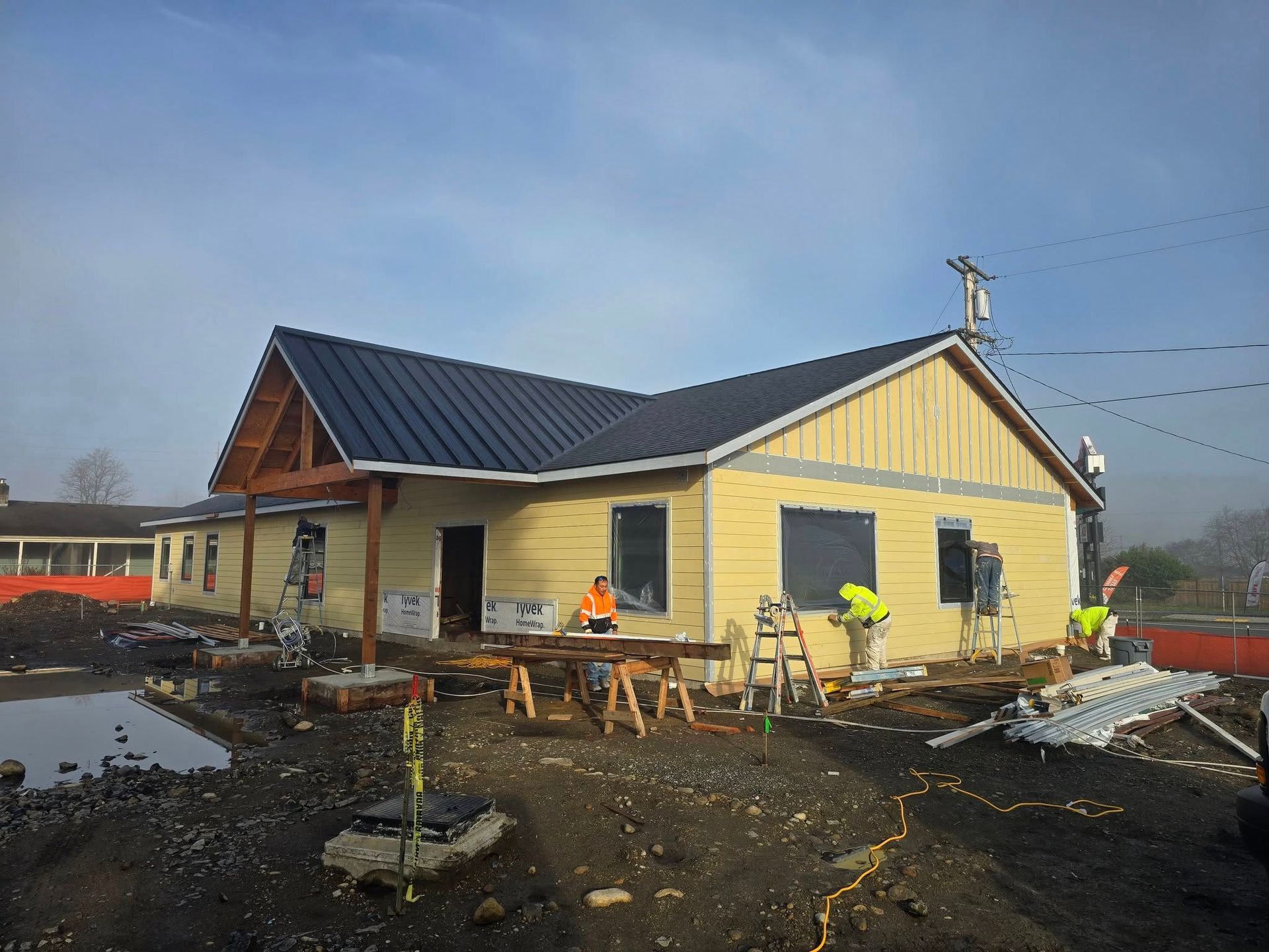Construction of a yellow building with dark roof, workers, and tools on a dirt lot.