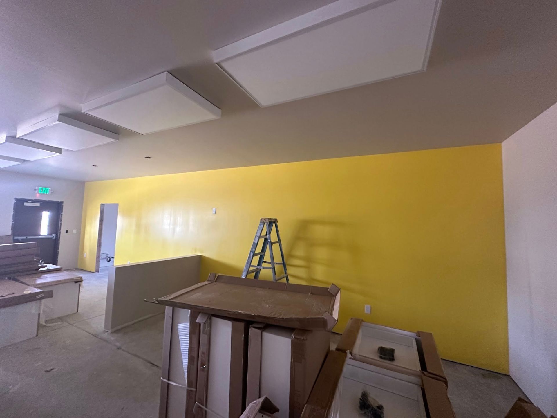 Interior view of a room under construction with a yellow wall, ladder, ceiling lights, and boxes.