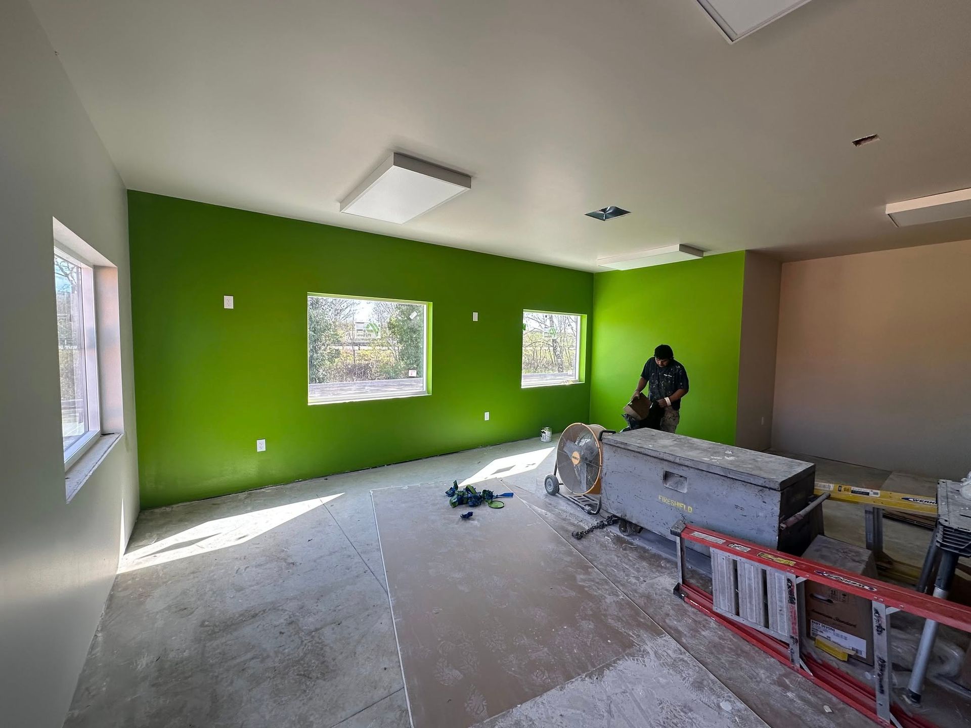 Green-walled room under construction; person near a toolbox. Unfinished floor, windows, and ceiling lights.
