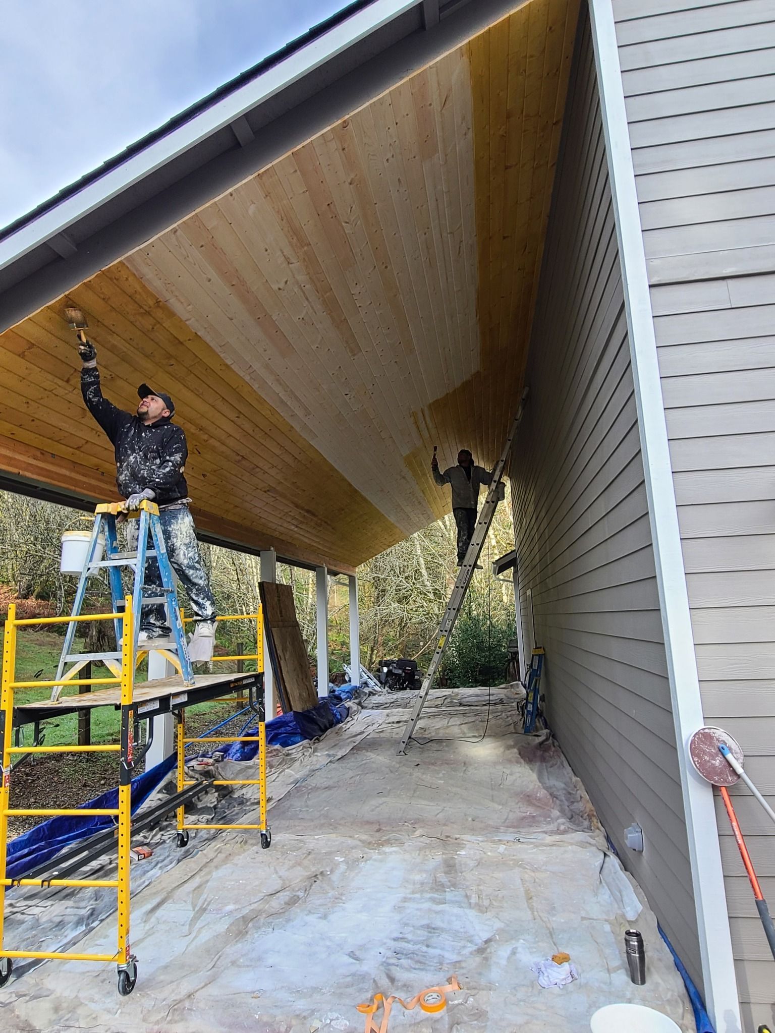 Two people painting a wooden ceiling under a house overhang. One on a ladder, the other on a taller support.