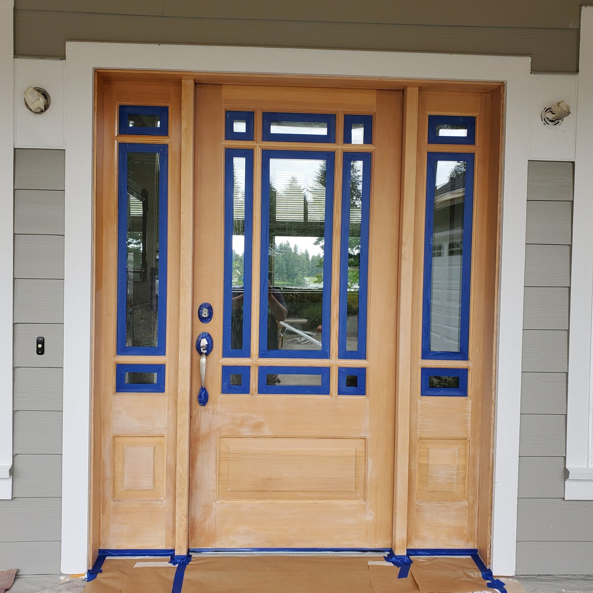 Wooden front door with glass panels taped off with blue painter's tape, ready for painting.