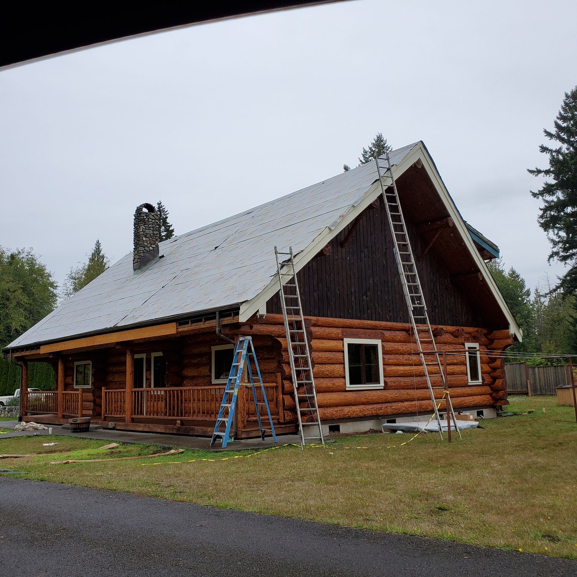 Log cabin under construction with ladders, metal roof, and brown logs against a cloudy sky.