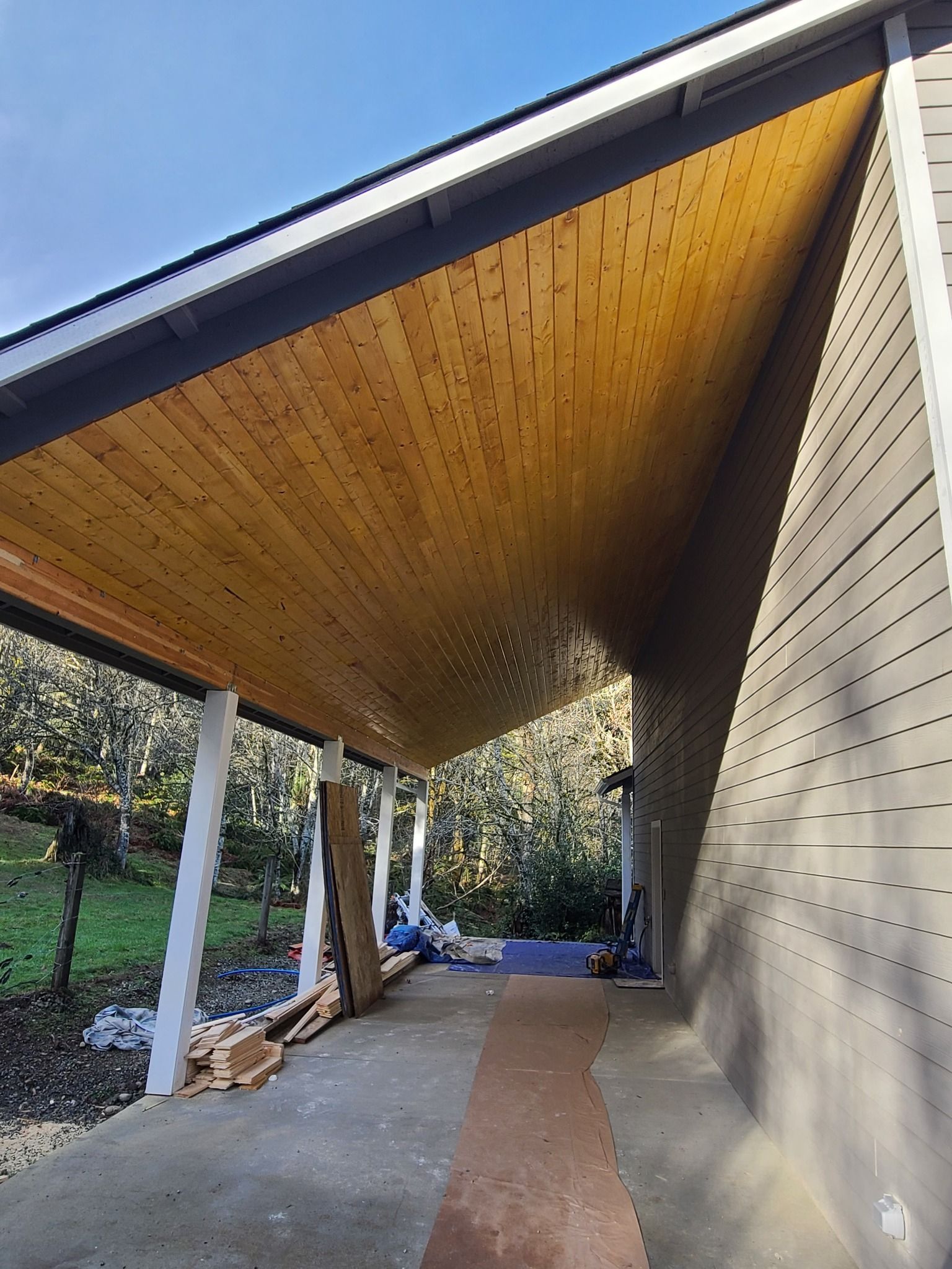 Covered porch with wooden ceiling and white columns, attached to a house with gray siding.