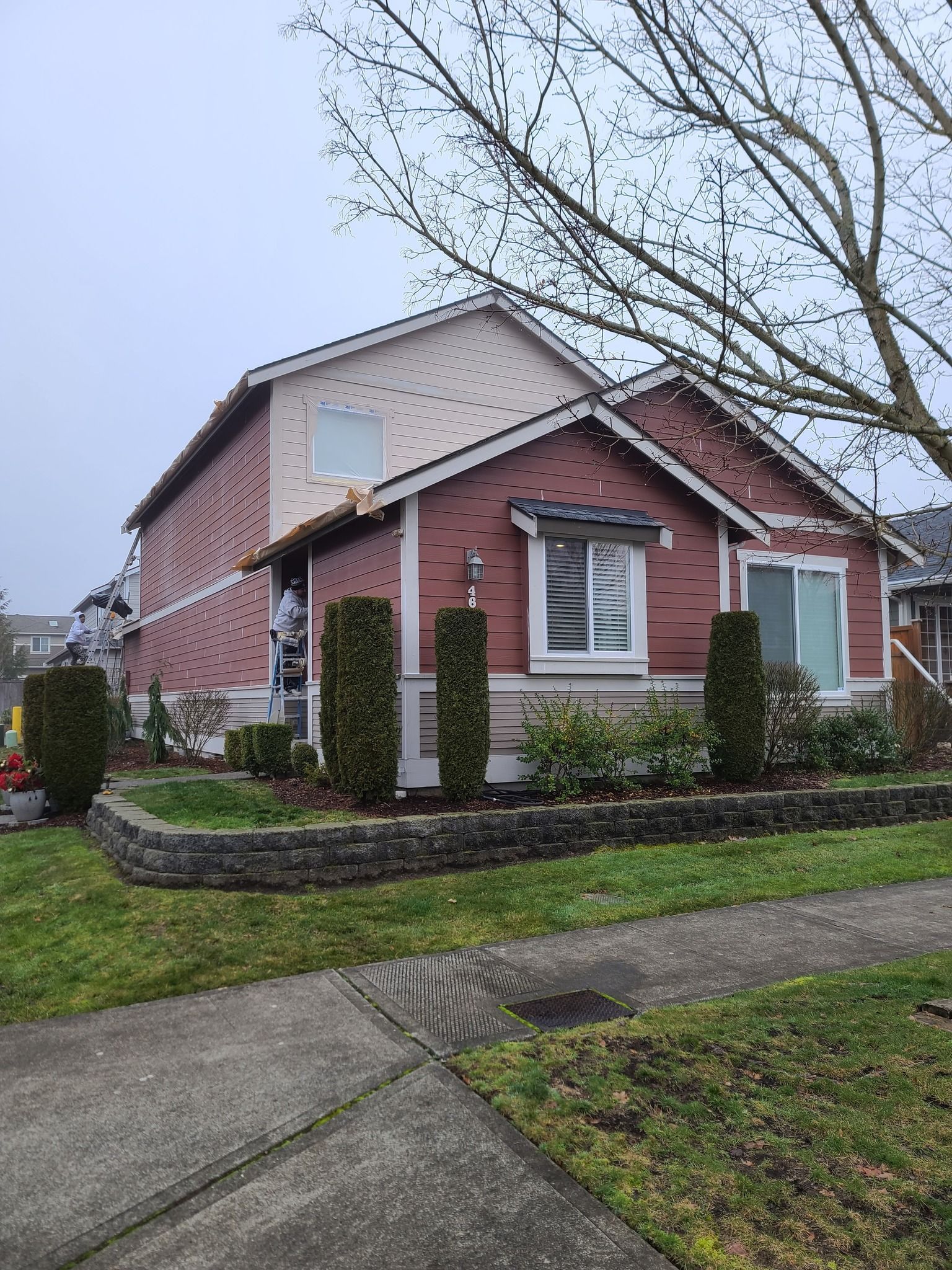 Two-story house with red siding and light tan roof on a cloudy day, featuring a low retaining wall with shrubs.