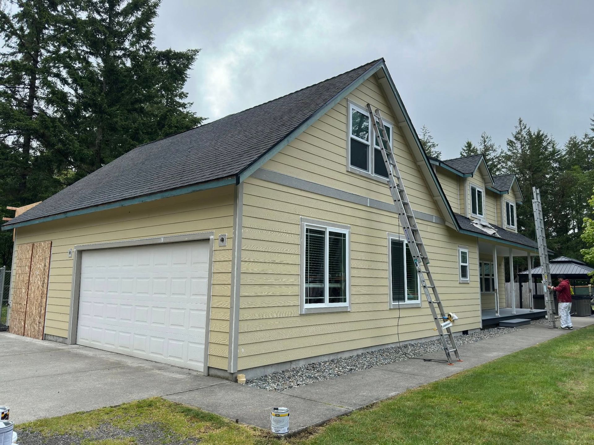 House exterior being painted, beige siding, white garage door, ladder, gray roof, overcast sky.