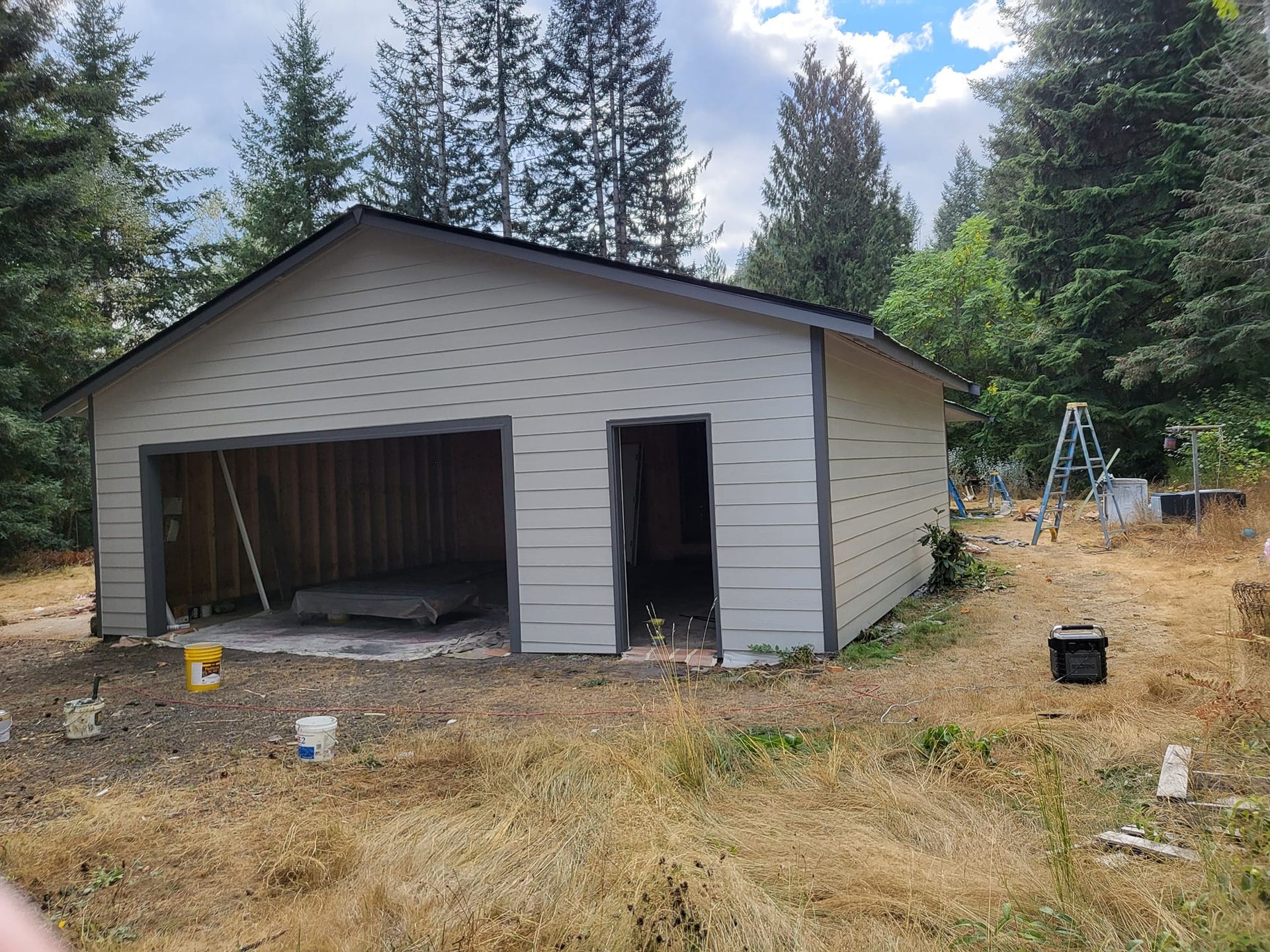 Garage with open bay door and man door, surrounded by dry grass and trees.