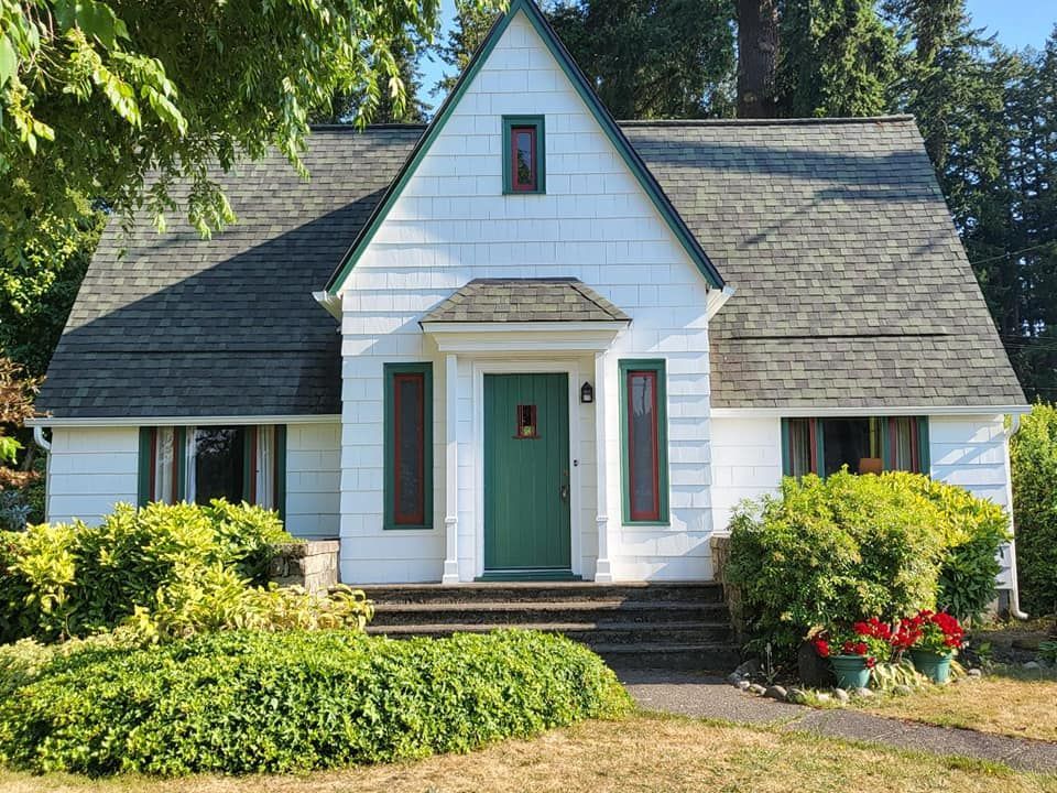 White cottage with green door and trim, gray roof, surrounded by green shrubs and a sunny lawn.