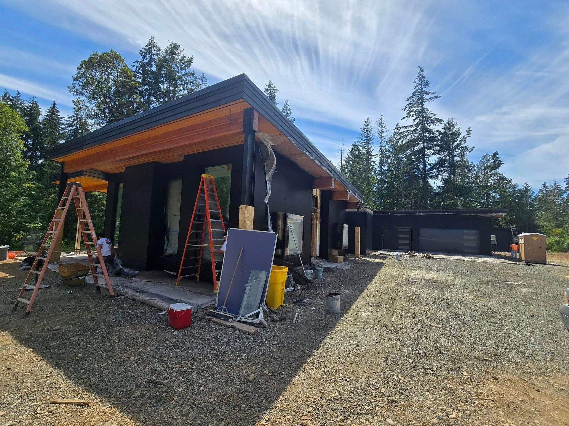 House under construction with black siding, wooden trim, and gravel driveway. Ladders and materials visible.