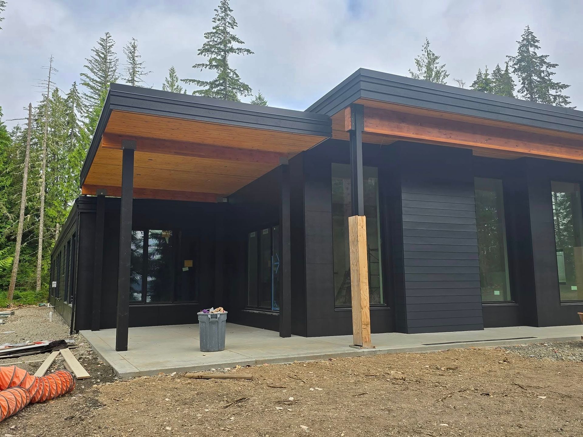 Black modern house with wood accents, covered patio, surrounded by trees and construction materials.