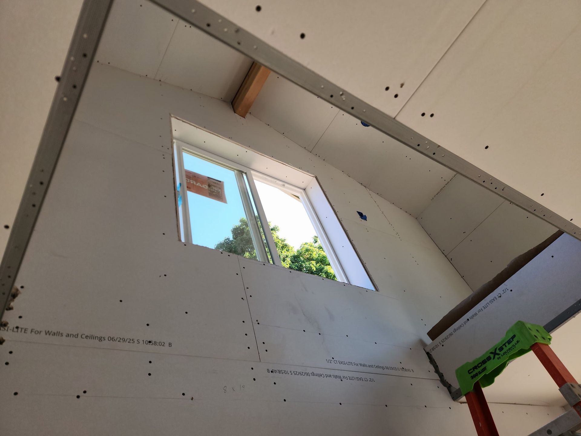 Interior view of drywall installation around a window; studs, screws, ladder visible.