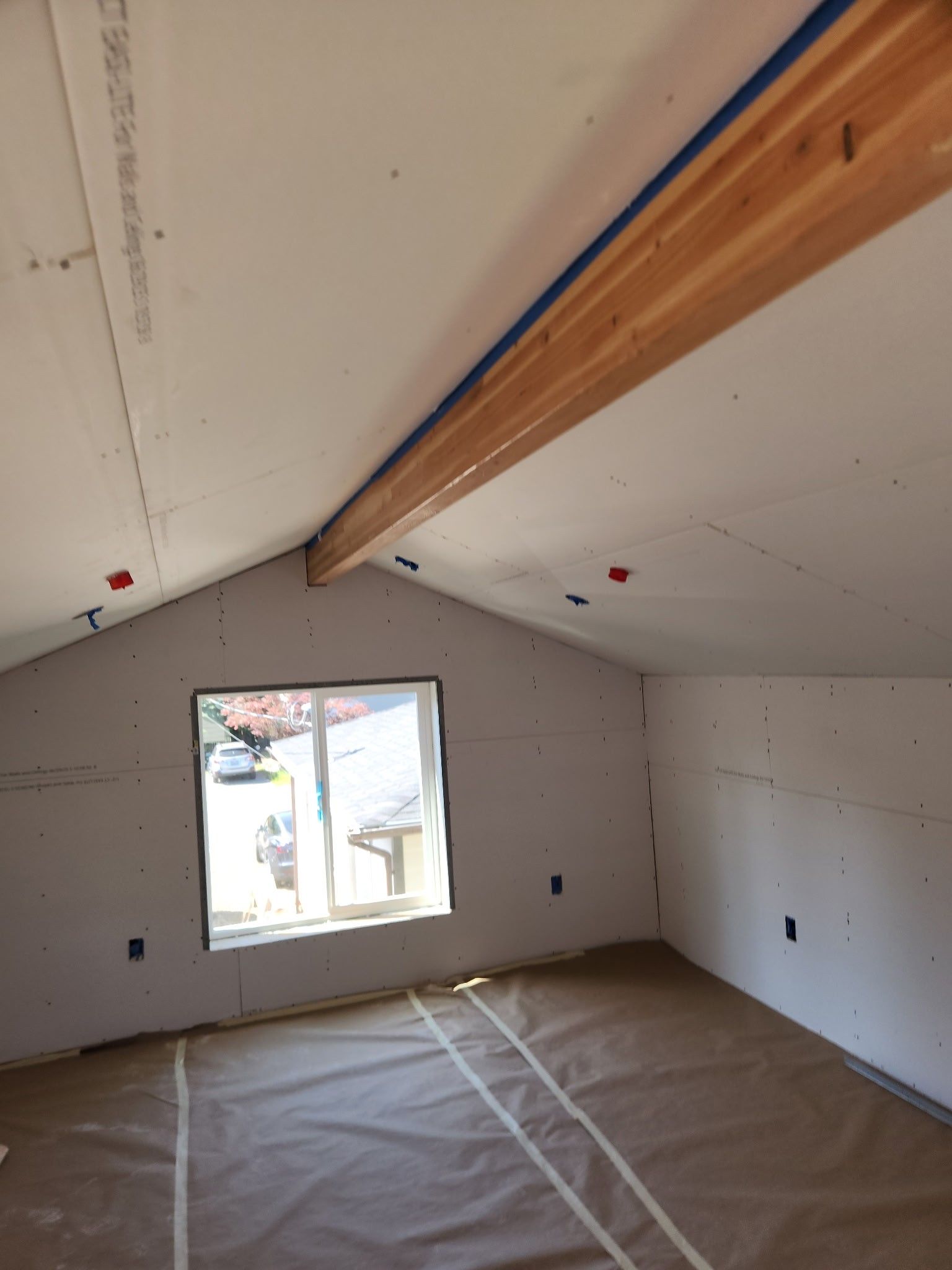 Interior of a room under construction with a sloped ceiling, window, and exposed wooden beam. Walls are covered with drywall.