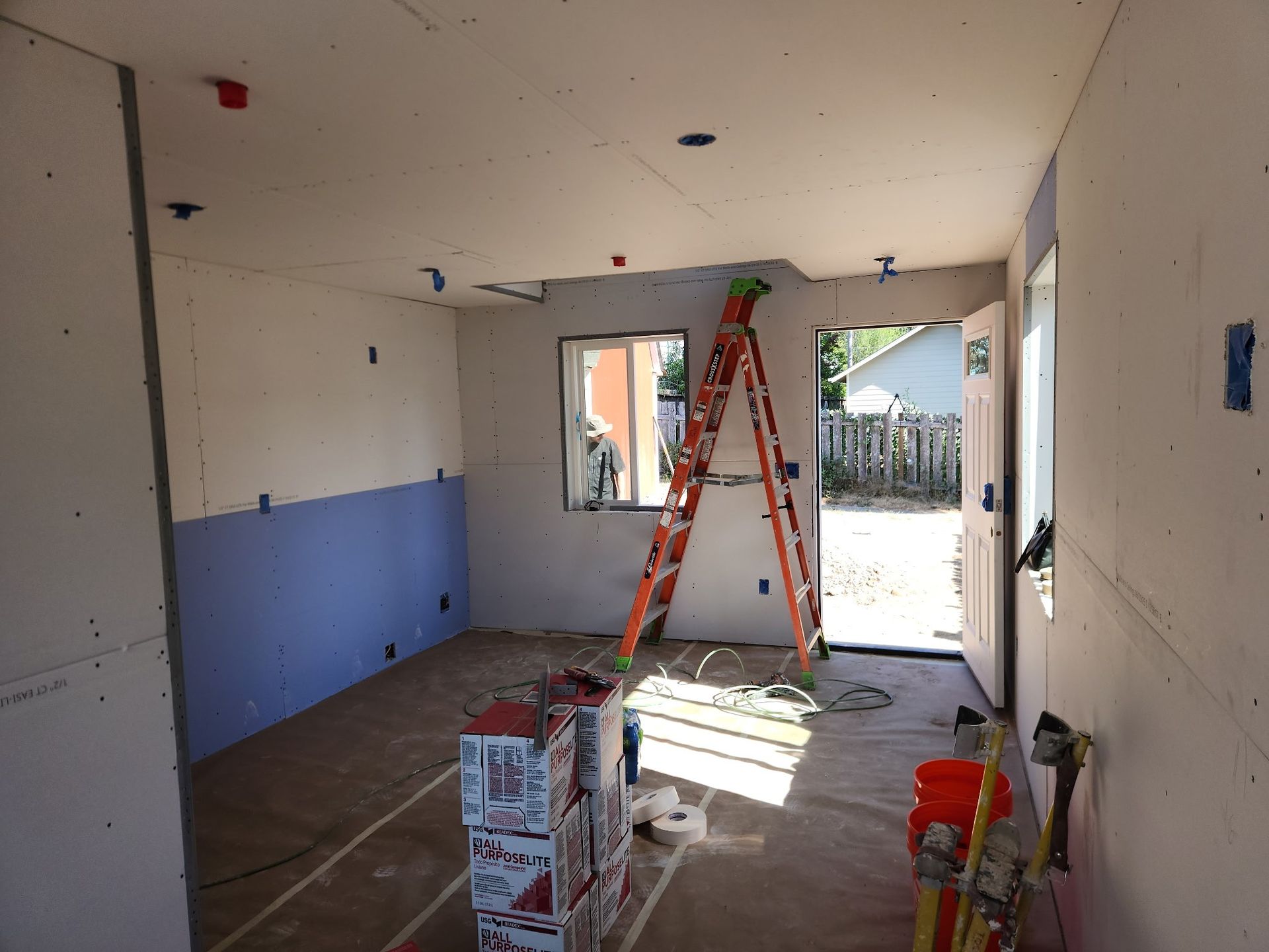 Interior of a room under construction with drywall and a ladder. Blue and white walls, door, window.