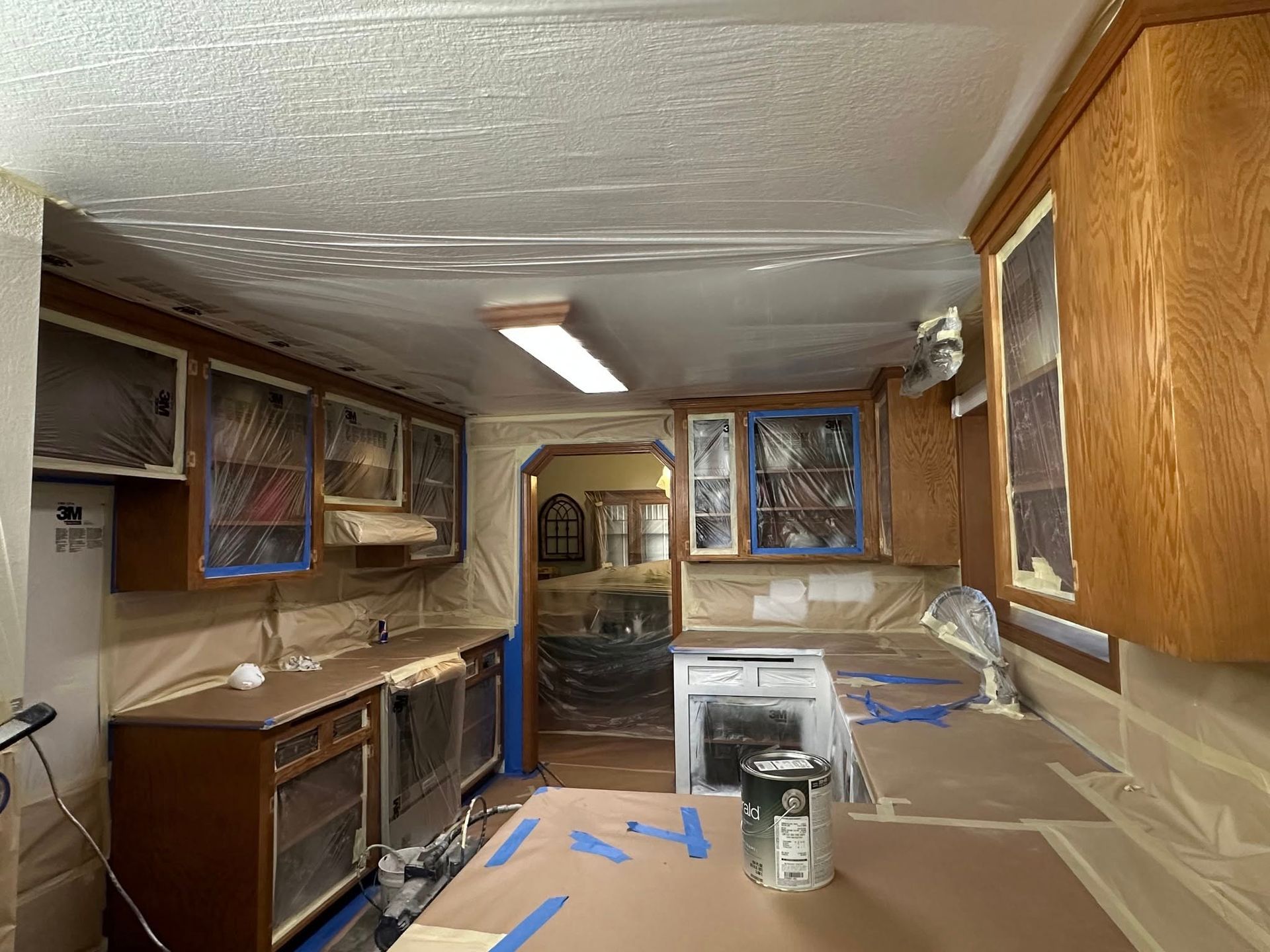 Kitchen renovation in progress: Cabinets and counters covered in plastic, taped for painting. Ceiling light fixture visible.