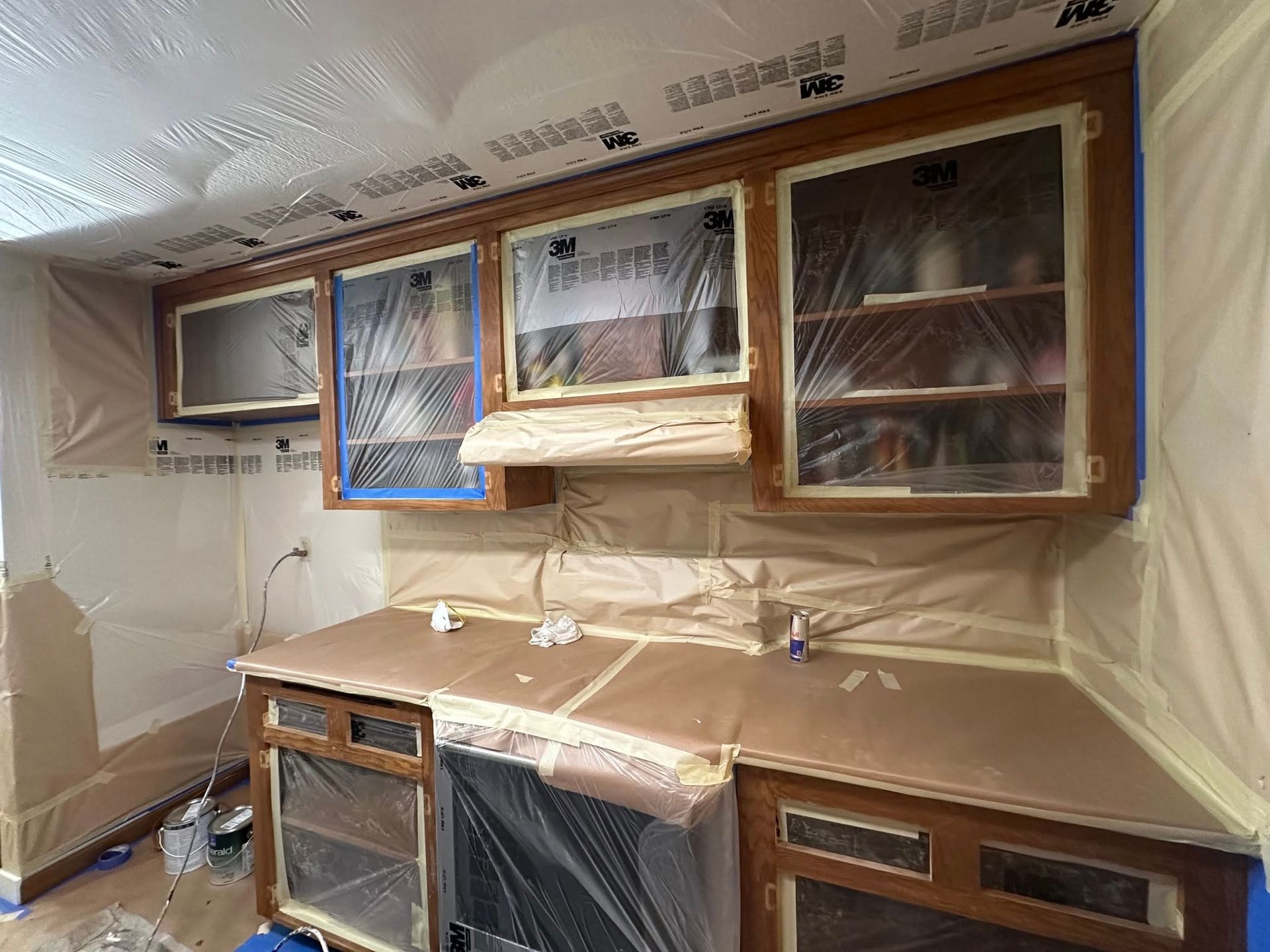 Kitchen cabinets prepped for painting: taped-off countertops, plastic-covered appliances and shelves.