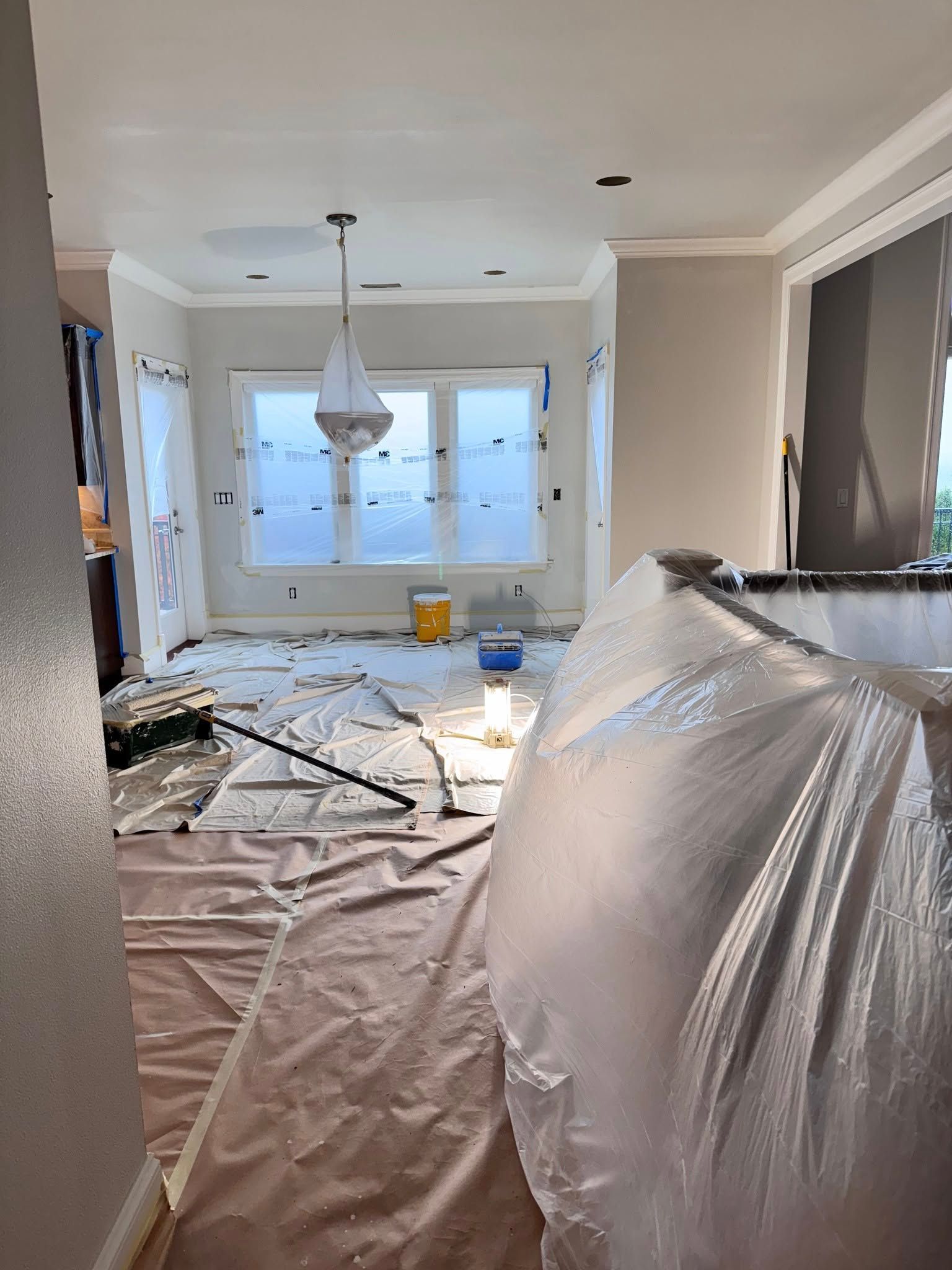 Room being painted, covered in protective plastic and drop cloths. Paint cans and a chandelier visible.