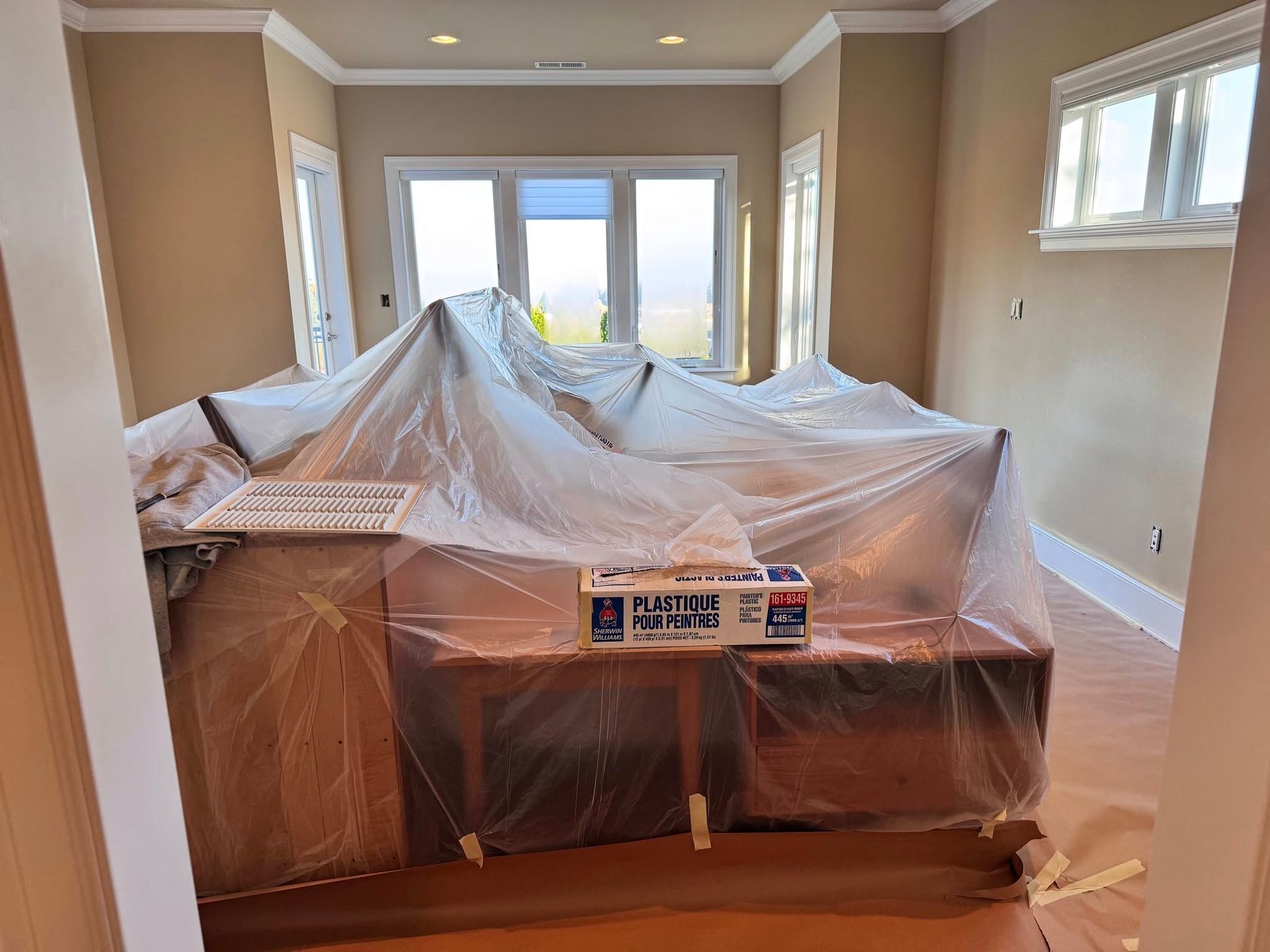 Room with a covered countertop under a large window, ready for renovations. Beige walls, wood flooring.
