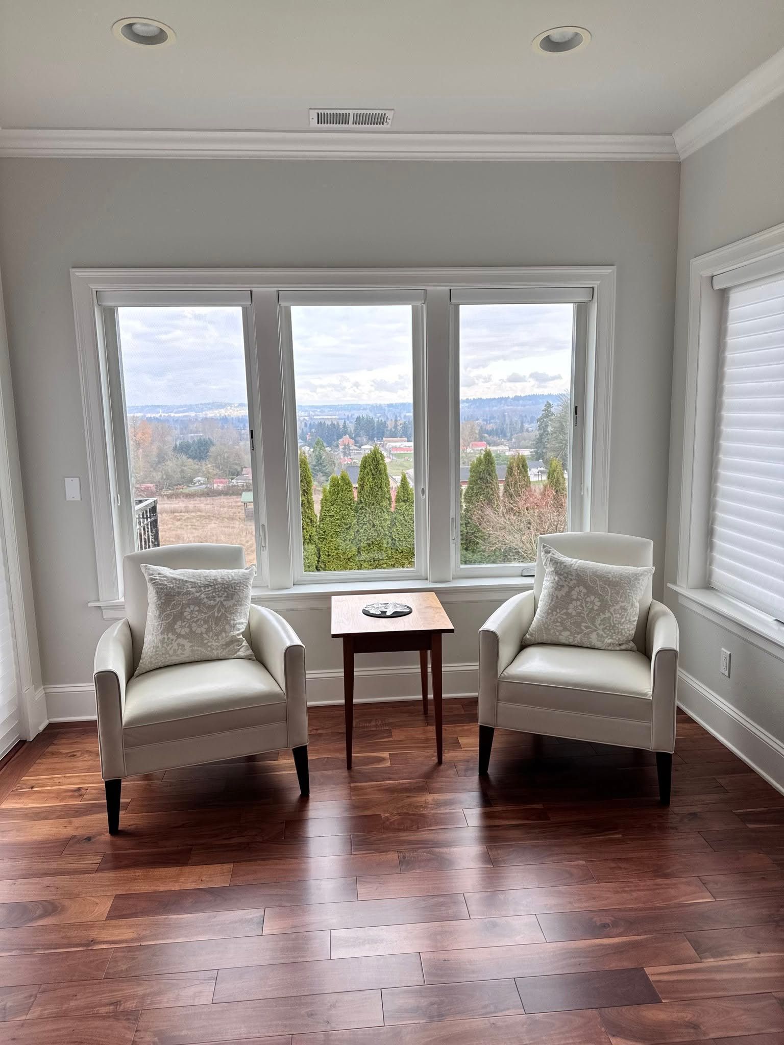 Two cream armchairs face a window with a city view; small wooden table between them, wooden floor.