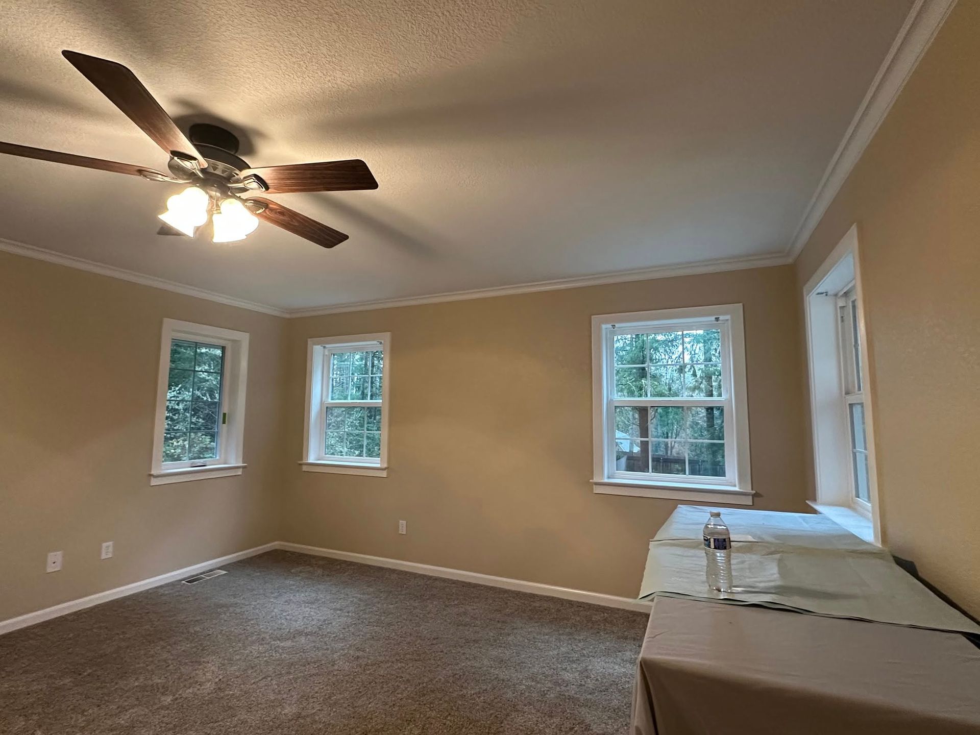 Empty room with three windows, a ceiling fan, beige walls, and brown carpet.