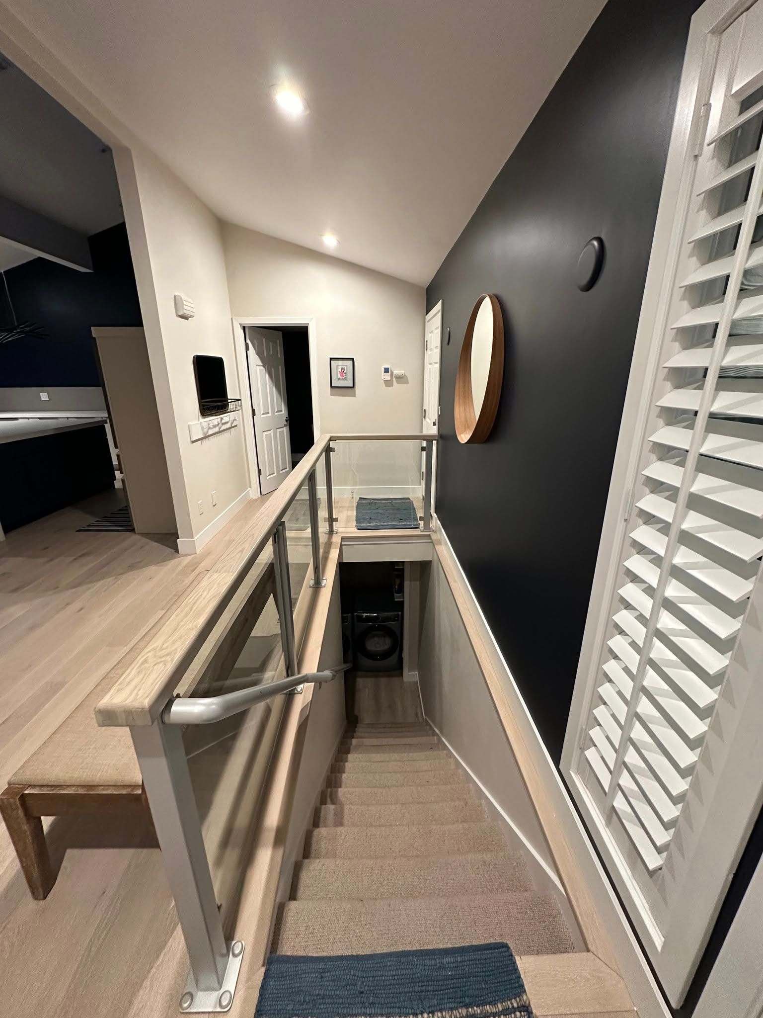Interior view of a staircase leading down to a laundry area; dark blue wall with mirror, wood flooring and bench.