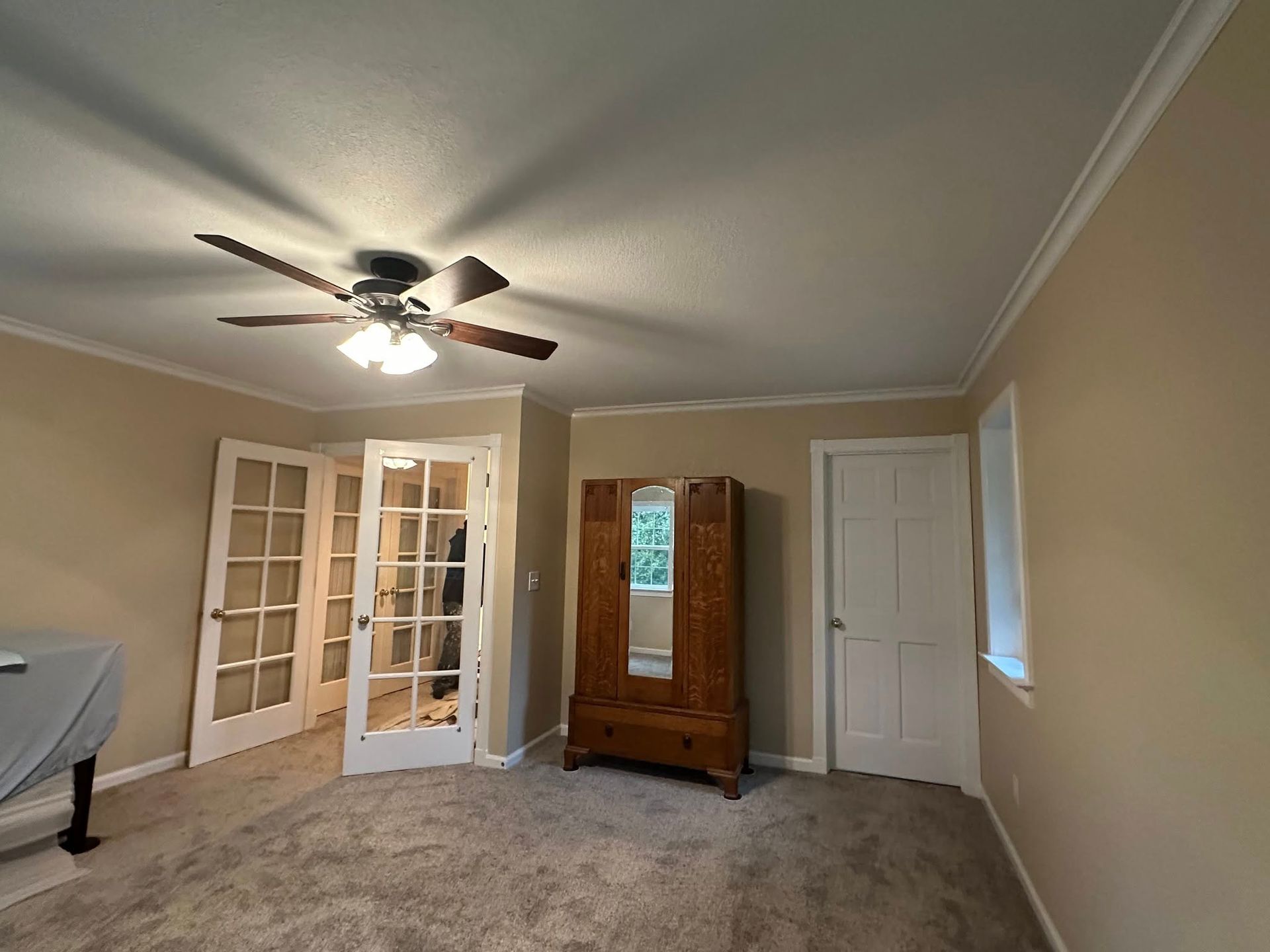 Room with French doors, an armoire, and a closed white door. Beige walls, gray carpet, and a ceiling fan.